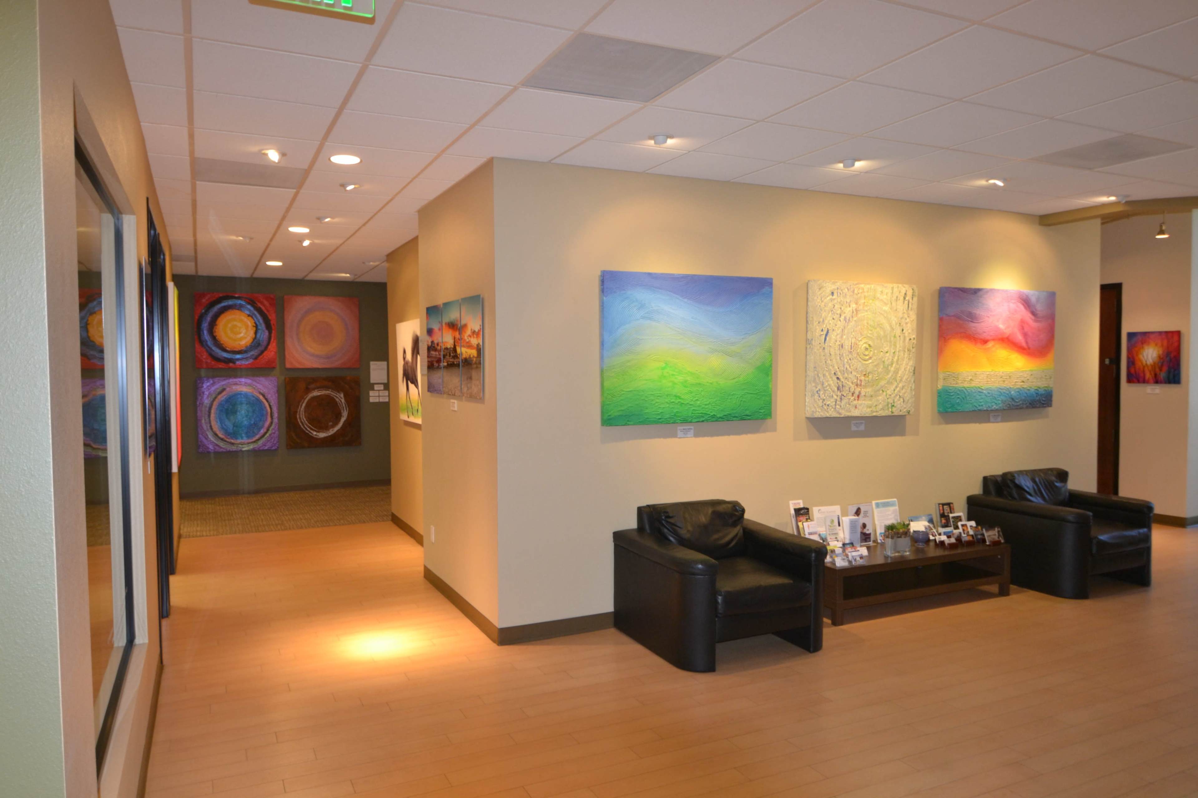 A well-lit hallway of an art gallery, featuring colorful abstract paintings on the walls and two black chairs placed against the wall.