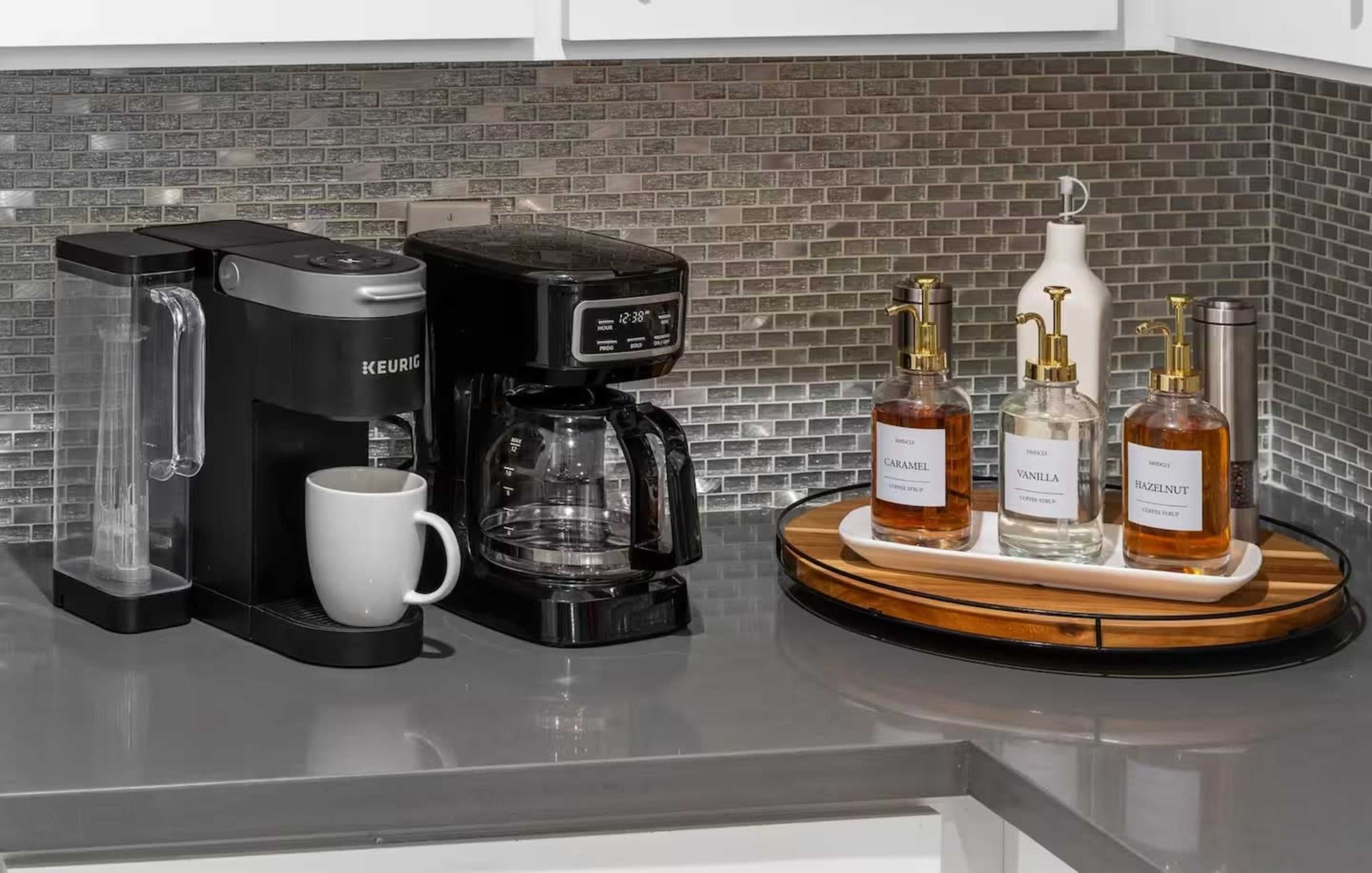 The image shows a kitchen countertop with a Keurig coffee maker, a traditional coffee brewer, a white mug, and a tray displaying bottles of liquid soap and lotion.