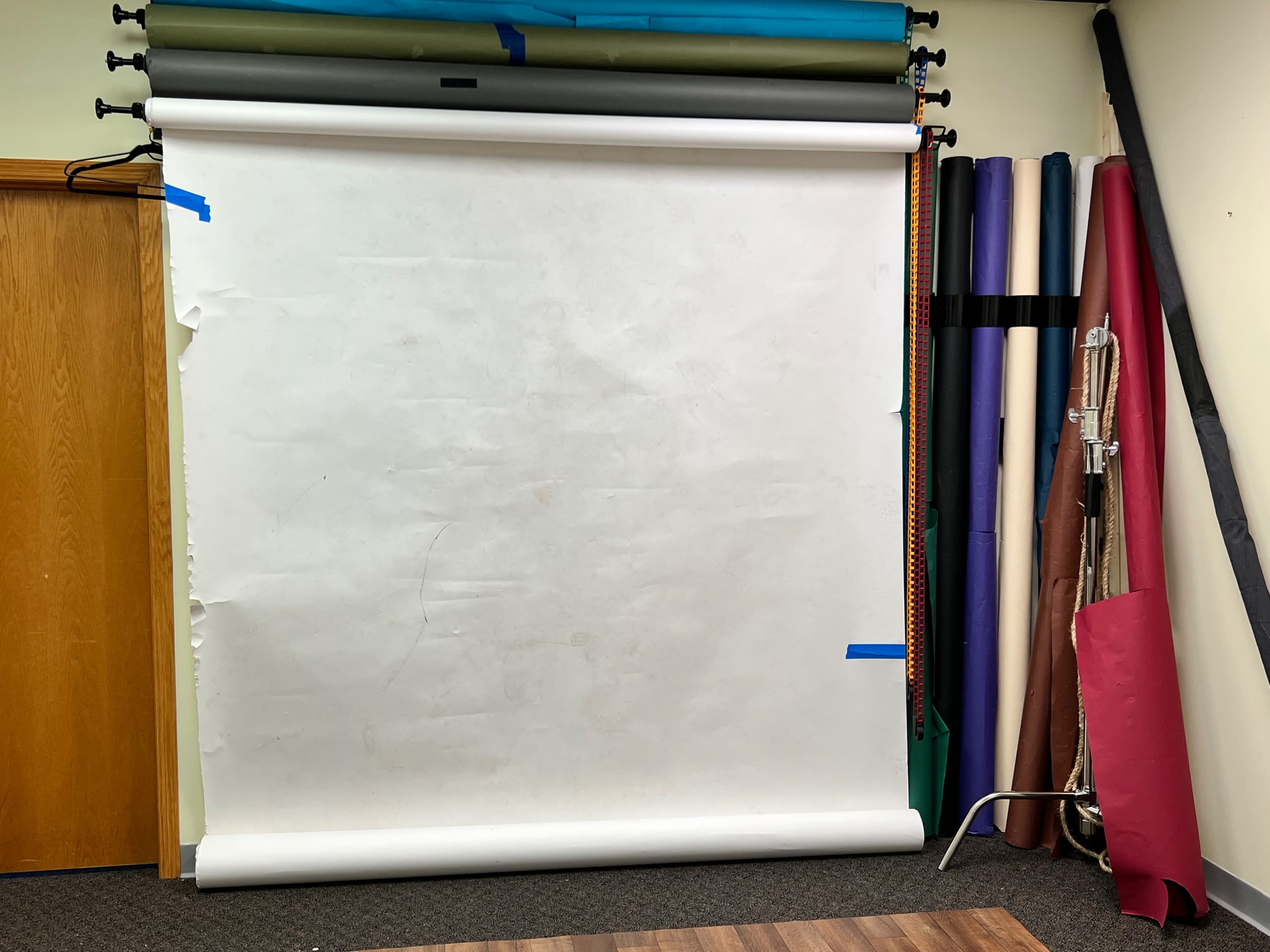 A photography backdrop setup with a large white roll-up backdrop and several rolls of colored paper positioned against a wall.