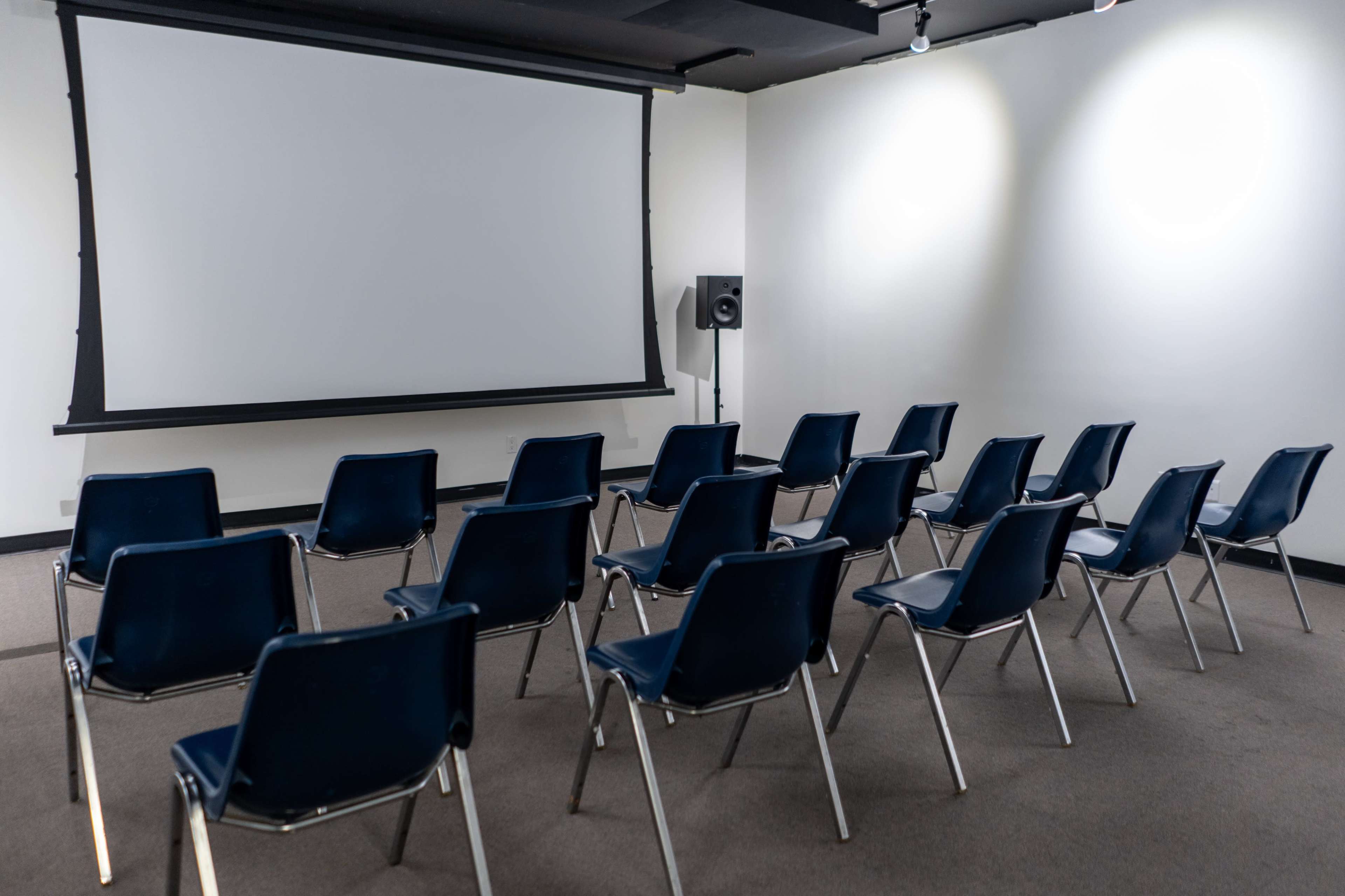 The image shows a small, empty room with rows of blue chairs facing a blank projector screen.