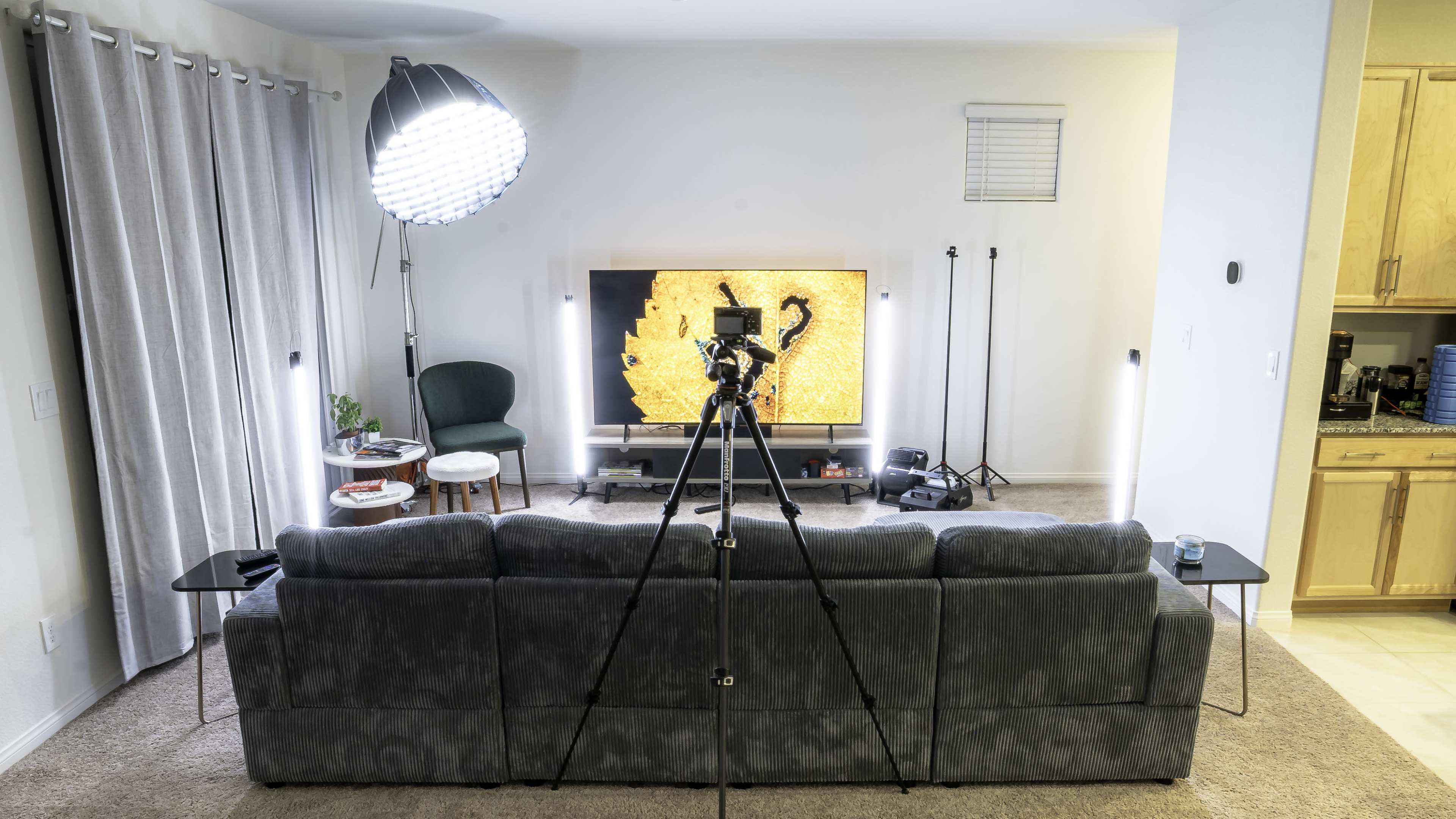 A camera is set up on a tripod facing a couch in a well-lit living room, with a large television displaying an image of a scorpion in the background.