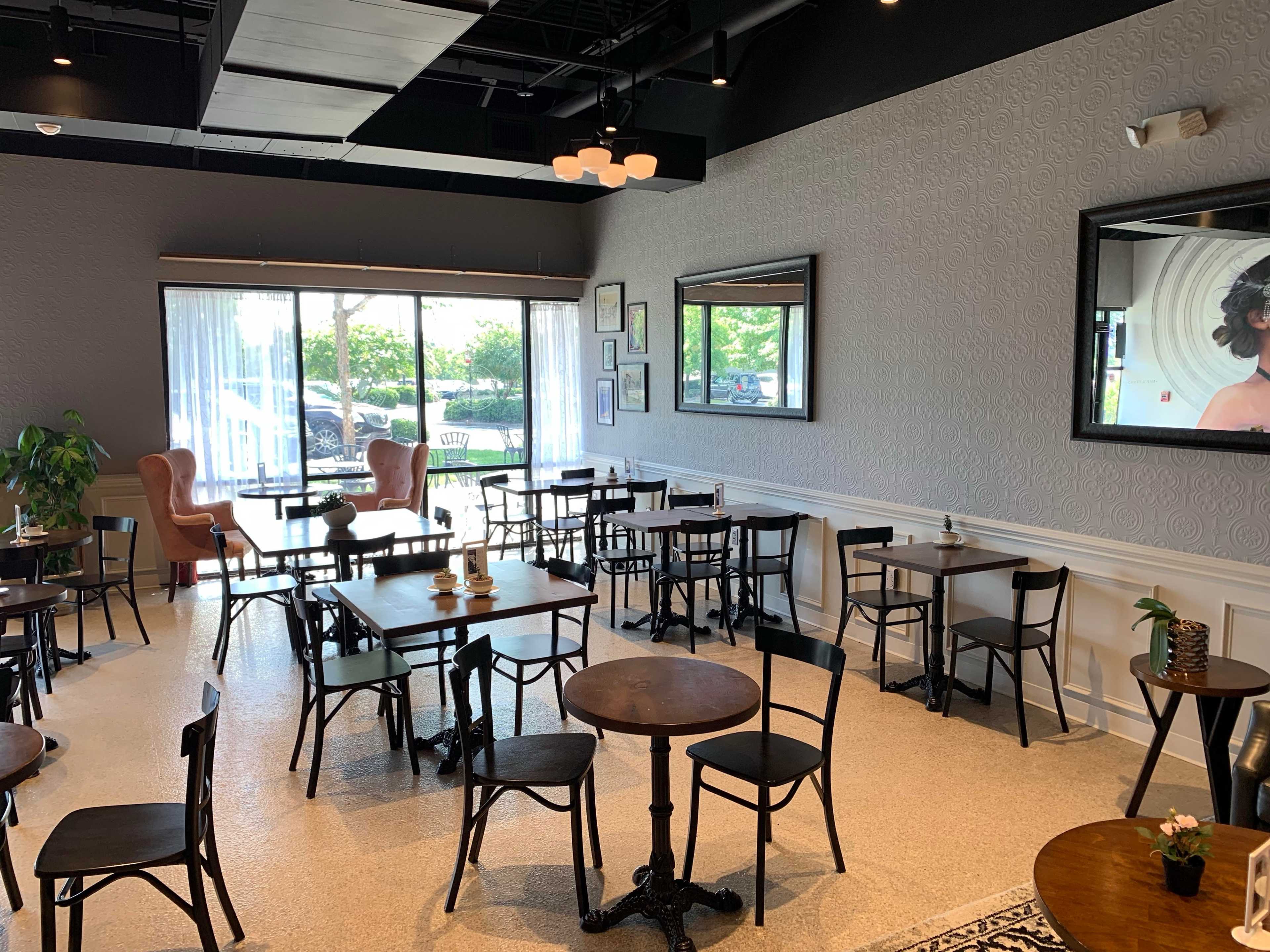 A spacious café interior with multiple tables and chairs, large windows allowing natural light, and a pair of armchairs in the corner.