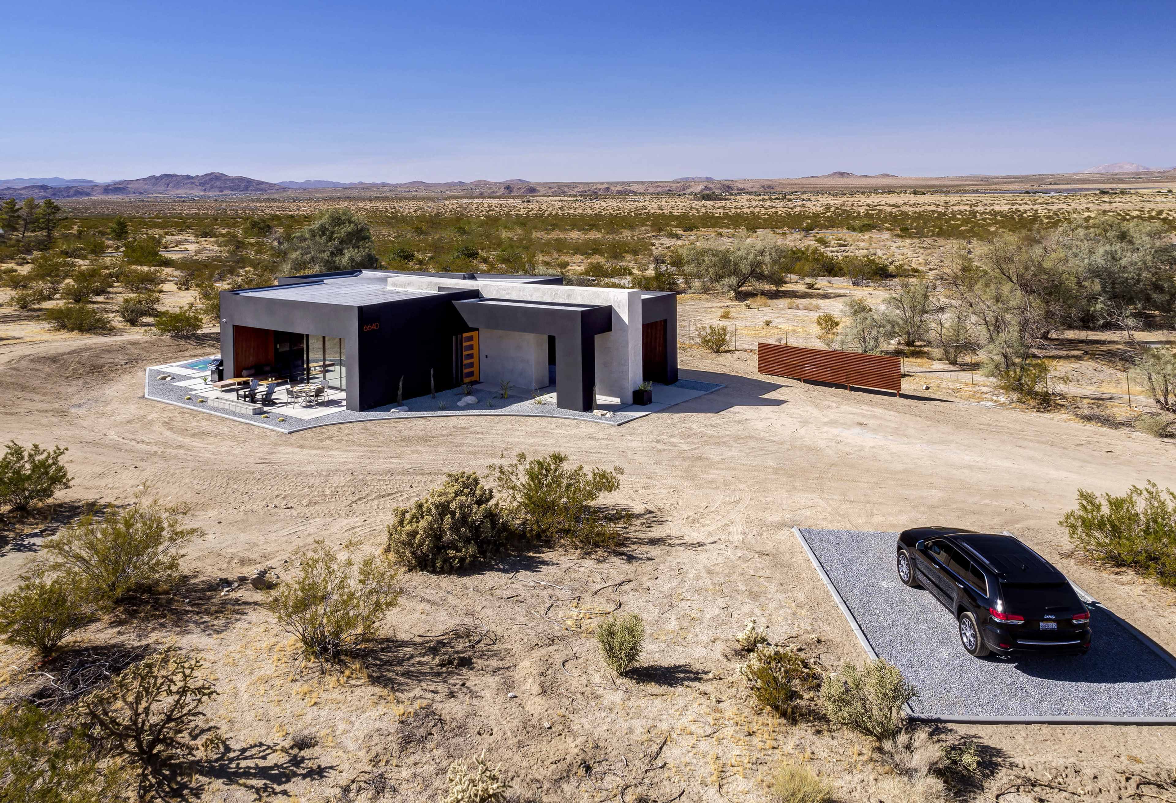 A modern, angular house sits in a barren desert landscape with sparse vegetation and a gravel driveway leading to a parked vehicle.