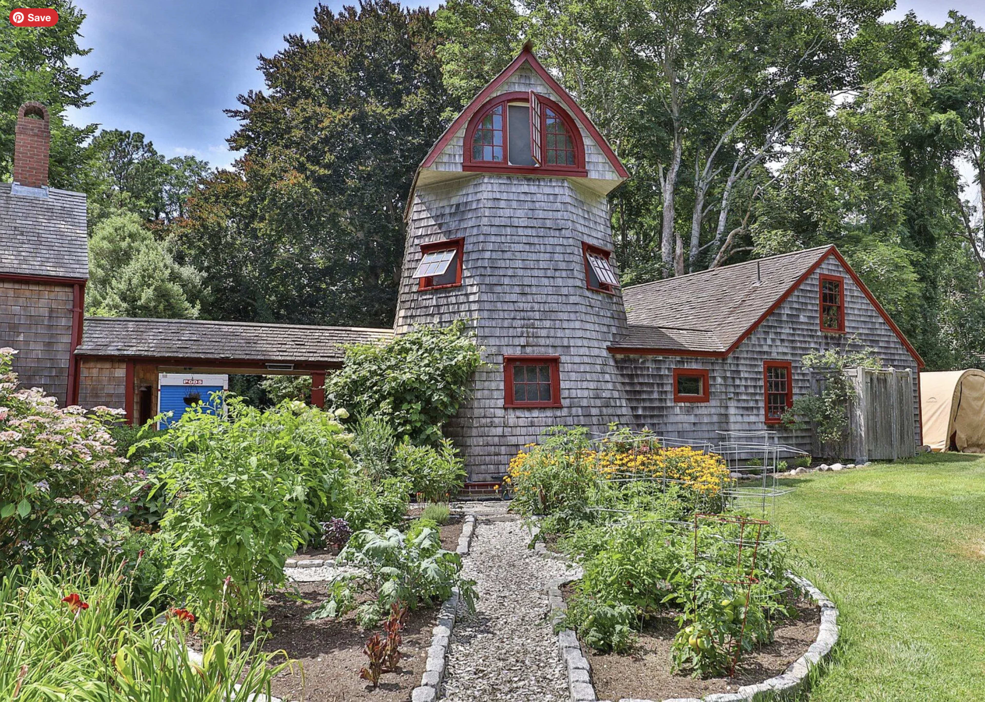 The image shows a two-story wooden house with a distinctive tower and a surrounding garden filled with various plants and flowers.