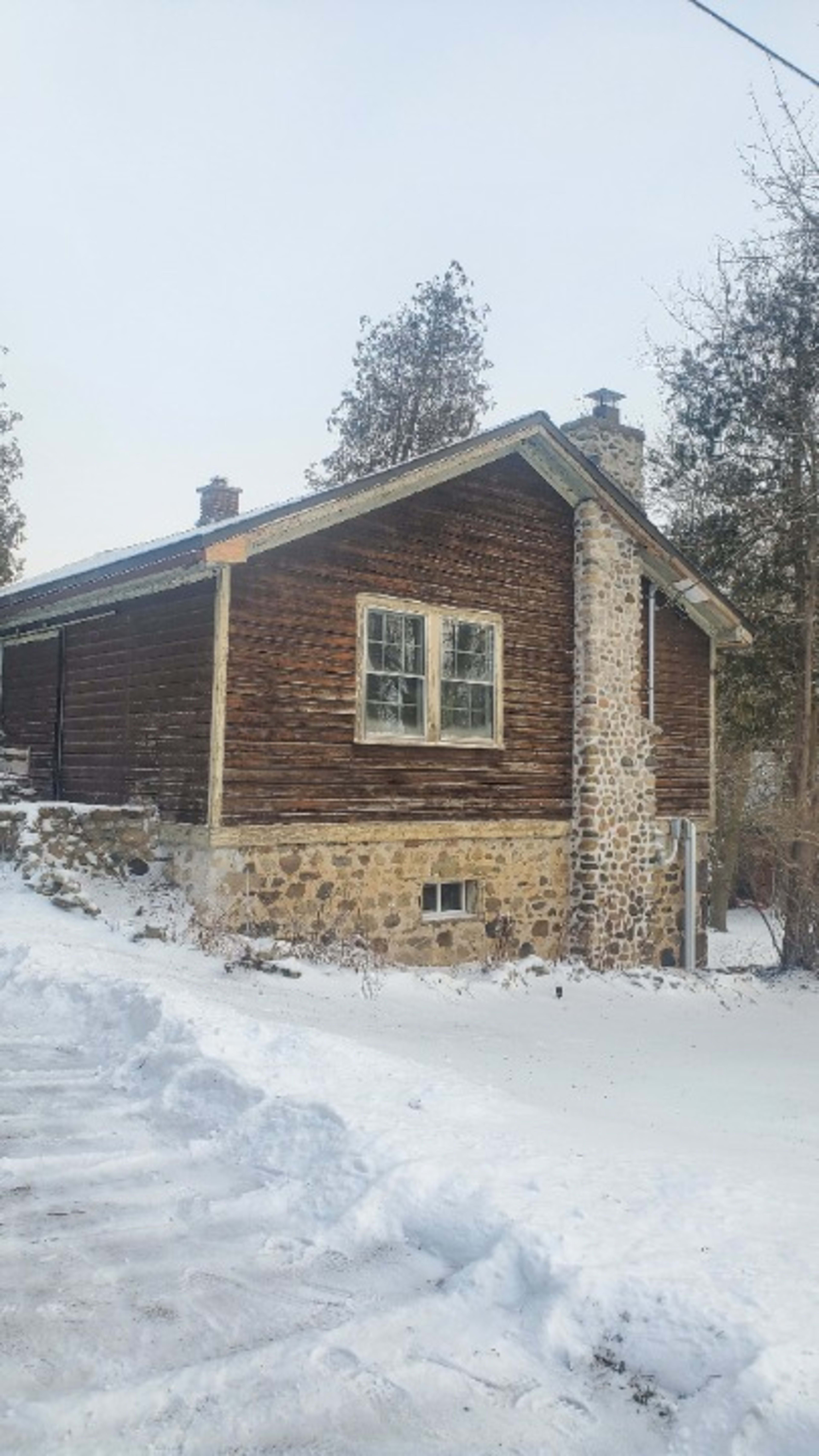 A two-story house with a wooden upper level and a stone lower level stands beside a snowy pathway.