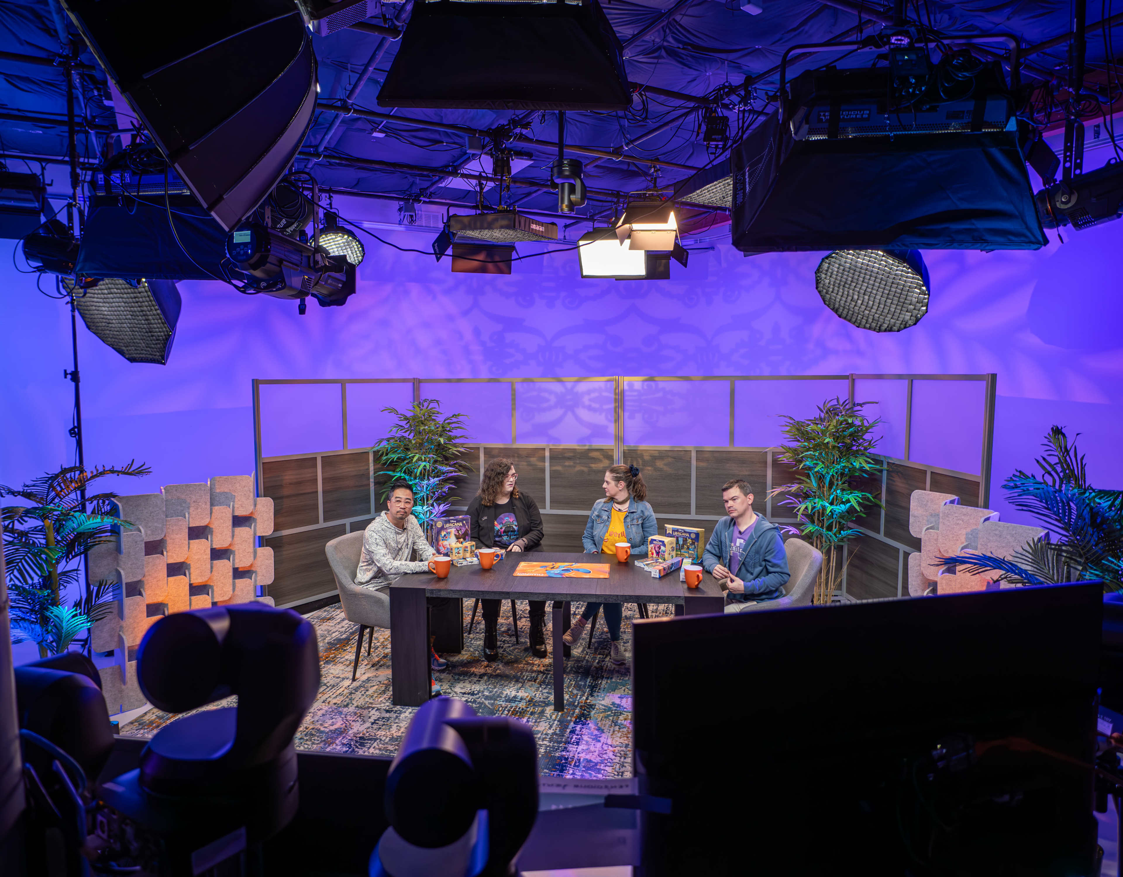 A group of four people is seated around a table in a brightly lit studio setting, surrounded by decorative plants and studio equipment.