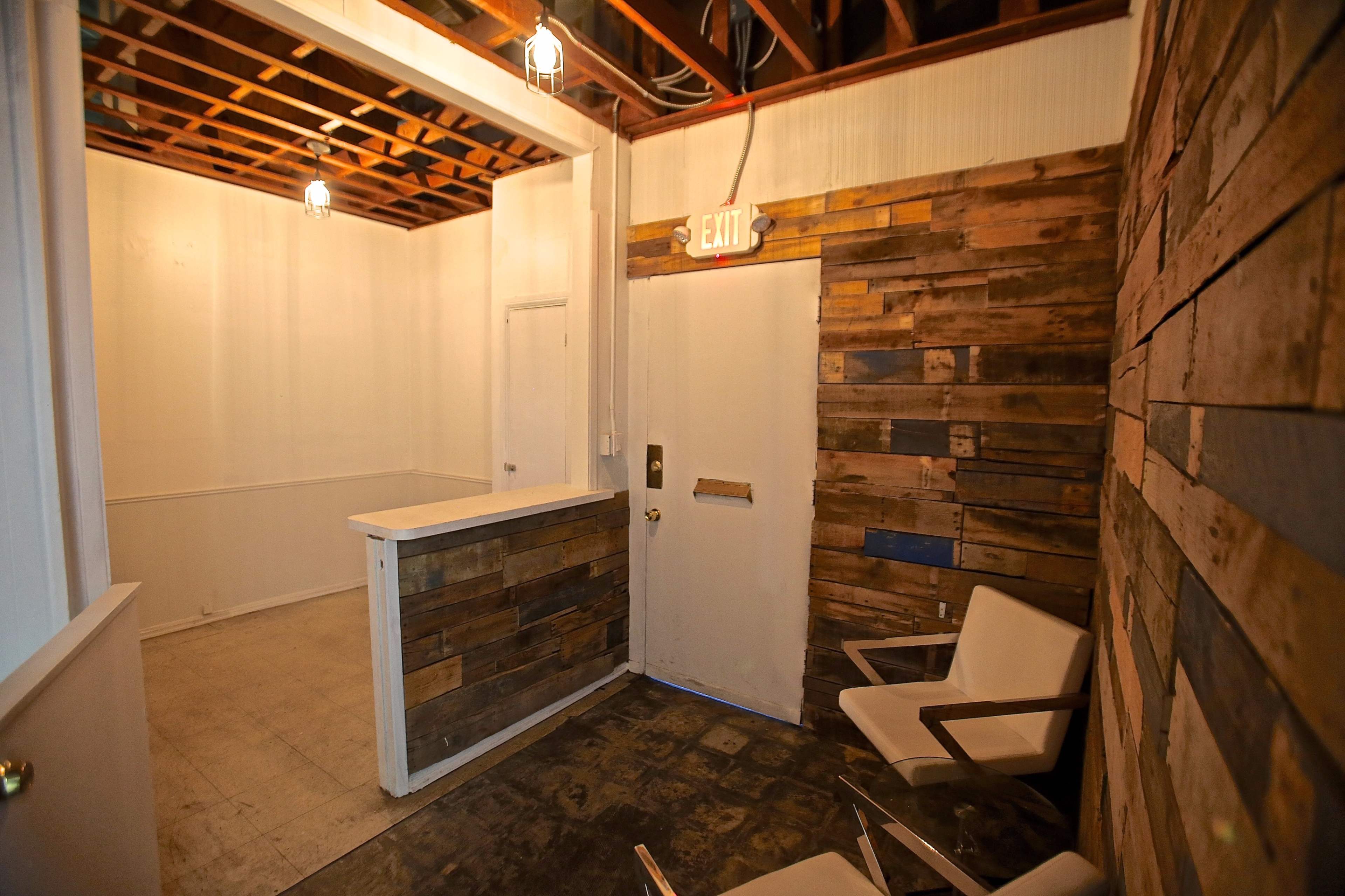 The image shows a small, minimally furnished room with wooden walls, a counter, a door, and a couple of chairs in the corner.