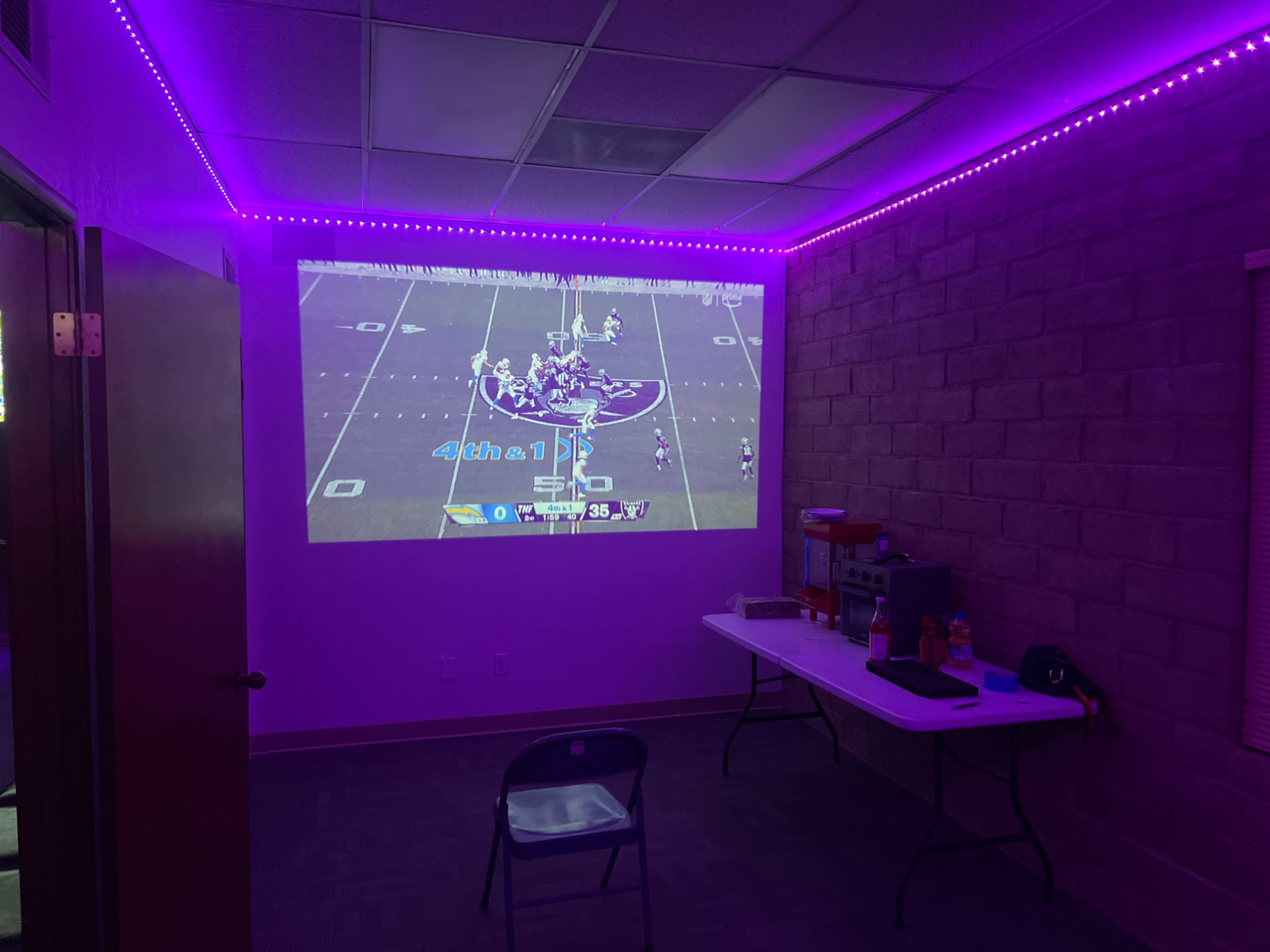 A football game is projected on a wall in a dimly lit room with purple LED lights.