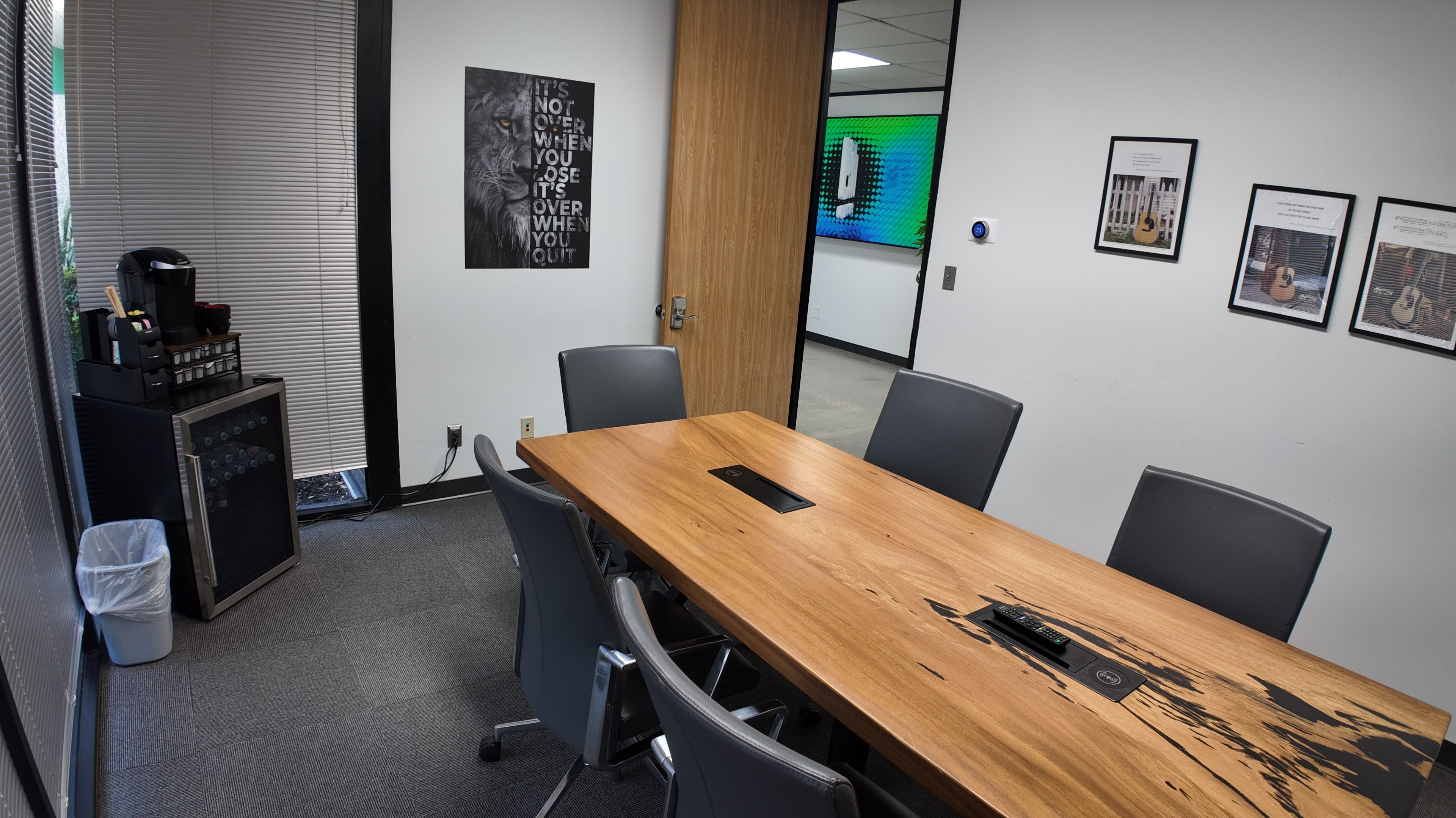 The image shows a modern conference room featuring a large wooden table with chairs, a beverage station, a small refrigerator, and framed artwork on the walls.