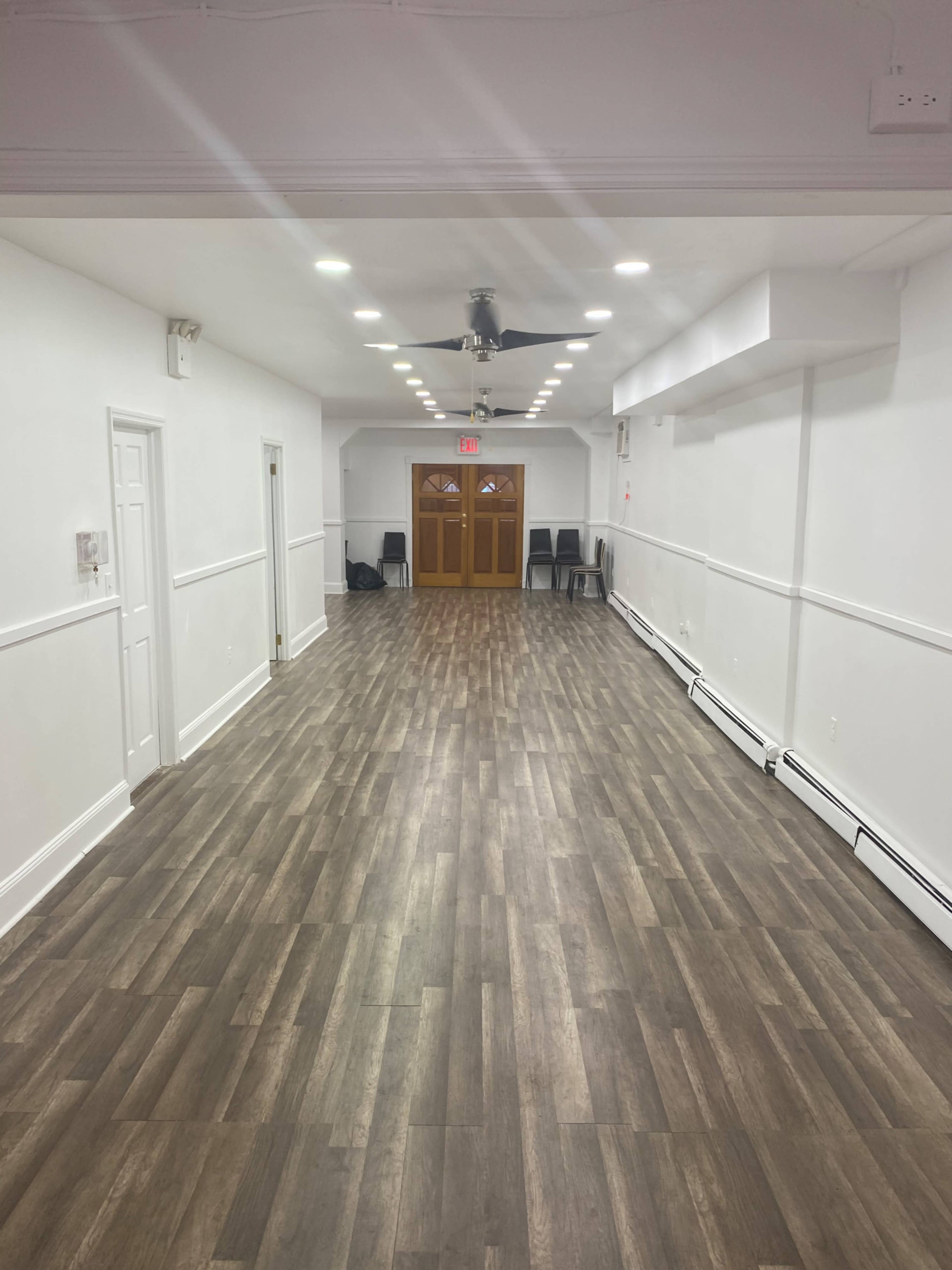 A long, empty room with wooden flooring, white walls, and a door at the far end leading outside.