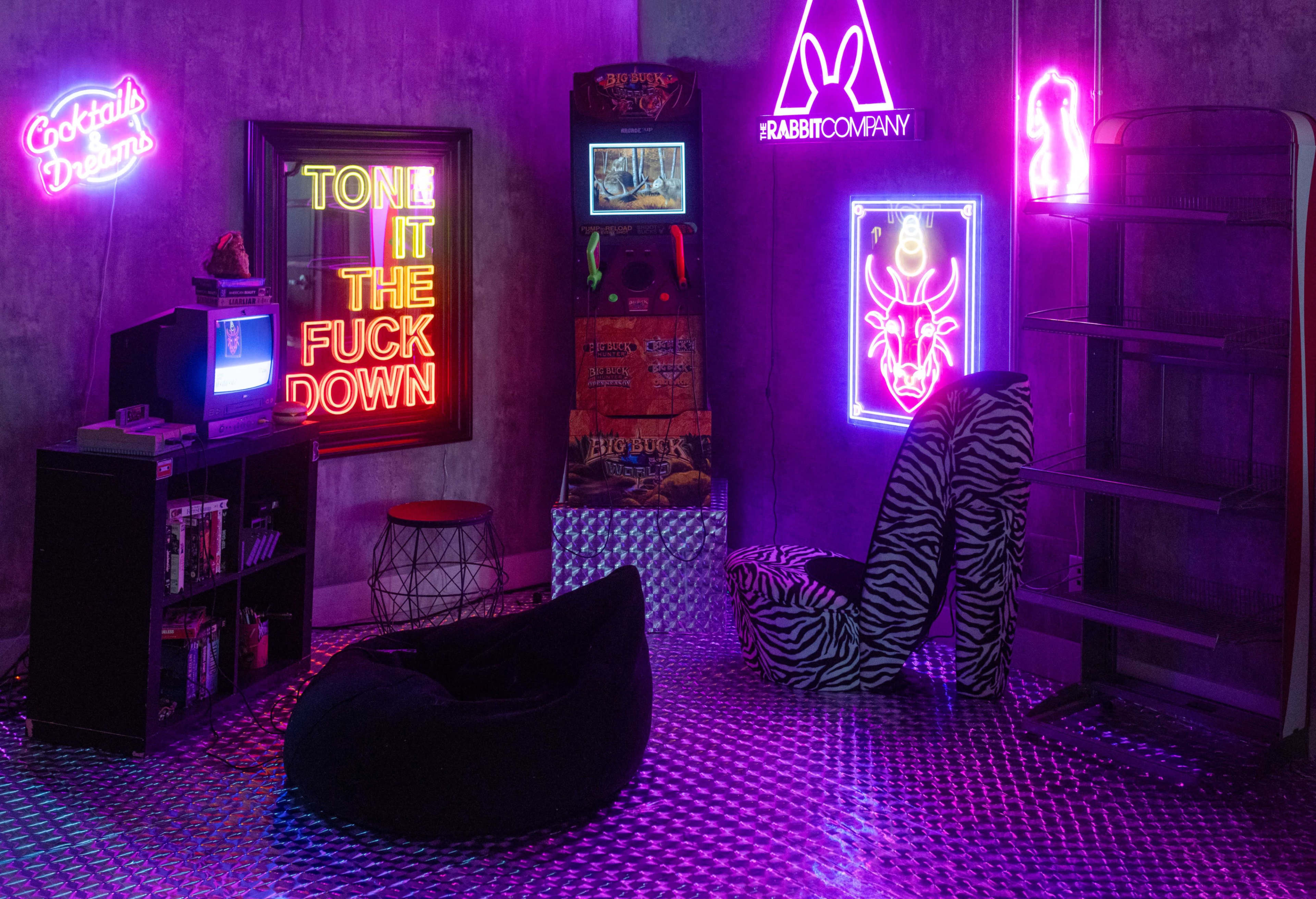 The image shows a retro-style room featuring neon signs, a vintage television, an arcade machine, and a bean bag chair on a reflective floor.