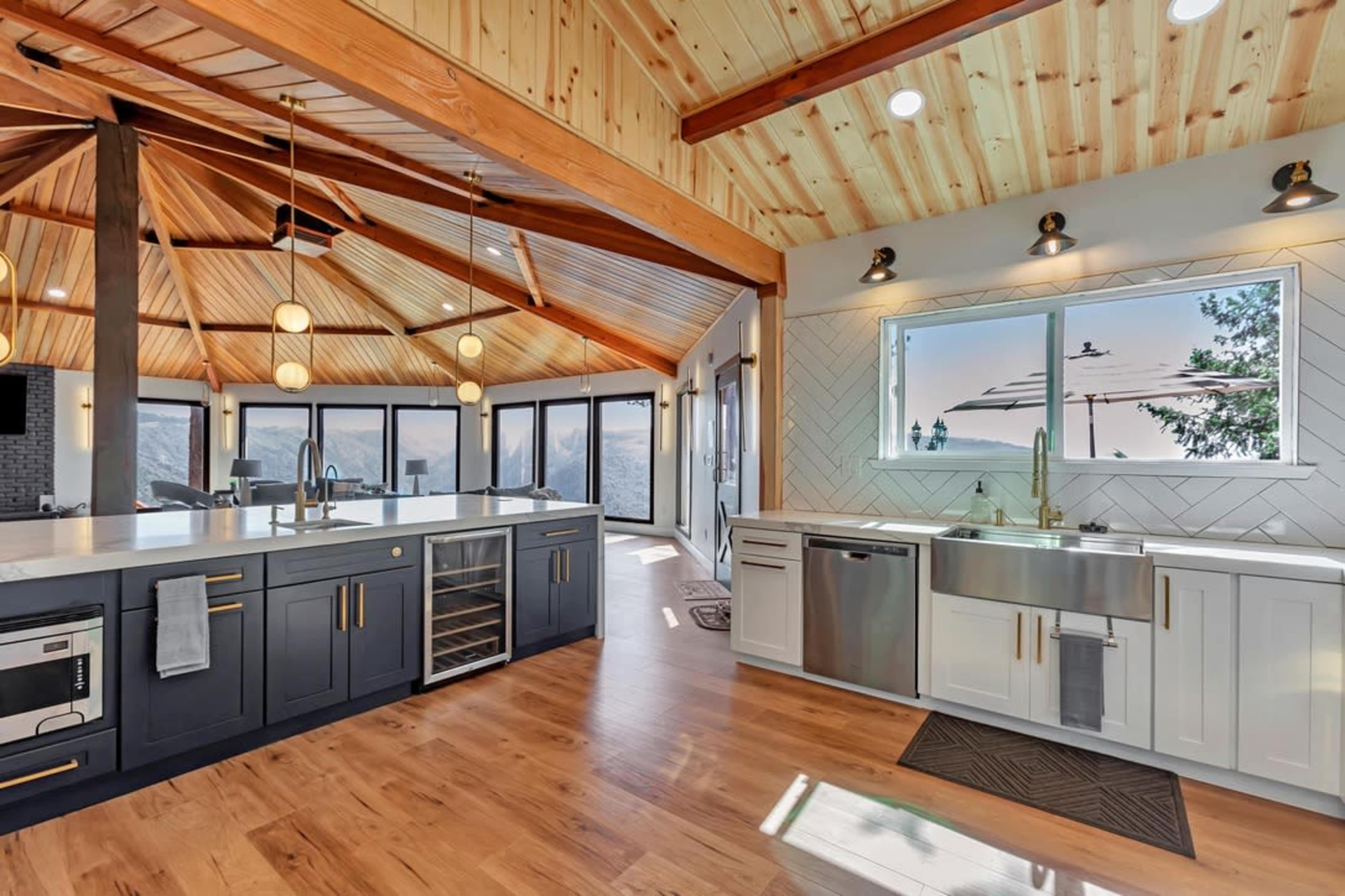 A modern kitchen with a dark blue island, white cabinetry, and large windows offering a view of the outdoors, all highlighted by a wood-paneled ceiling.