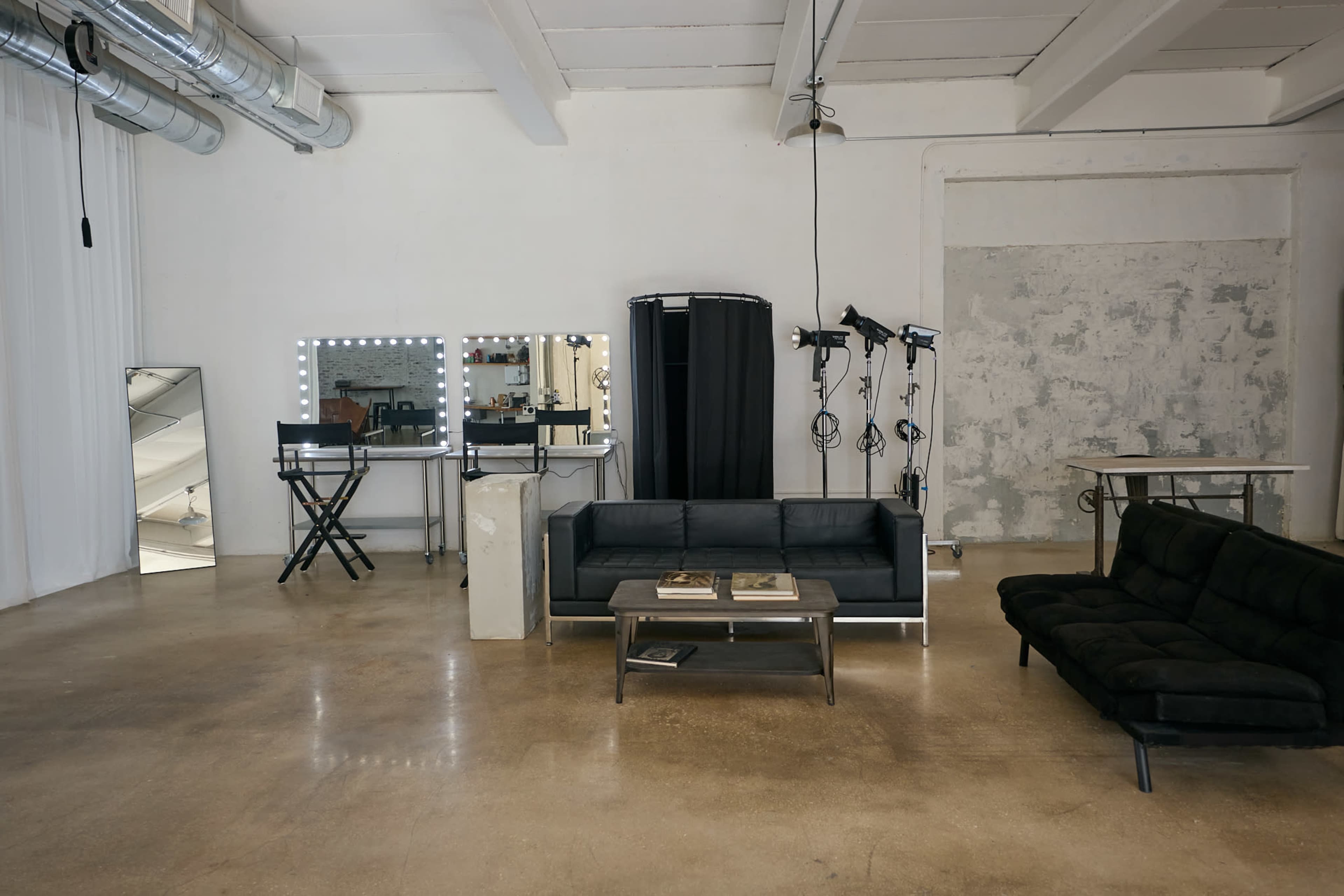 The image shows a spacious studio with a black sofa, a coffee table, tall mirrors, and studio lights arranged in a minimalist setting.