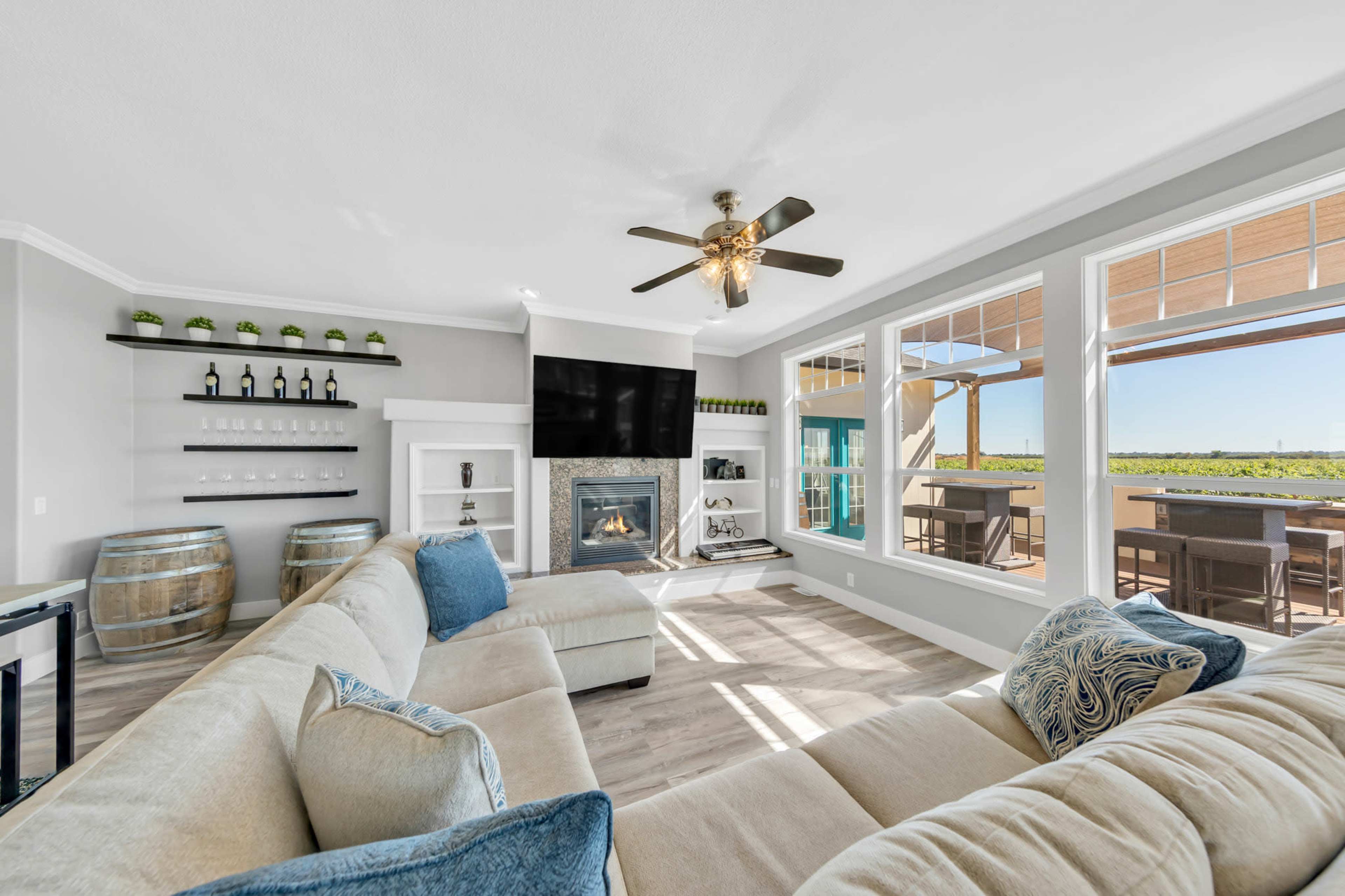 The living room features a large sectional sofa, a wall-mounted television above a fireplace, and large windows that offer views of the outdoors.