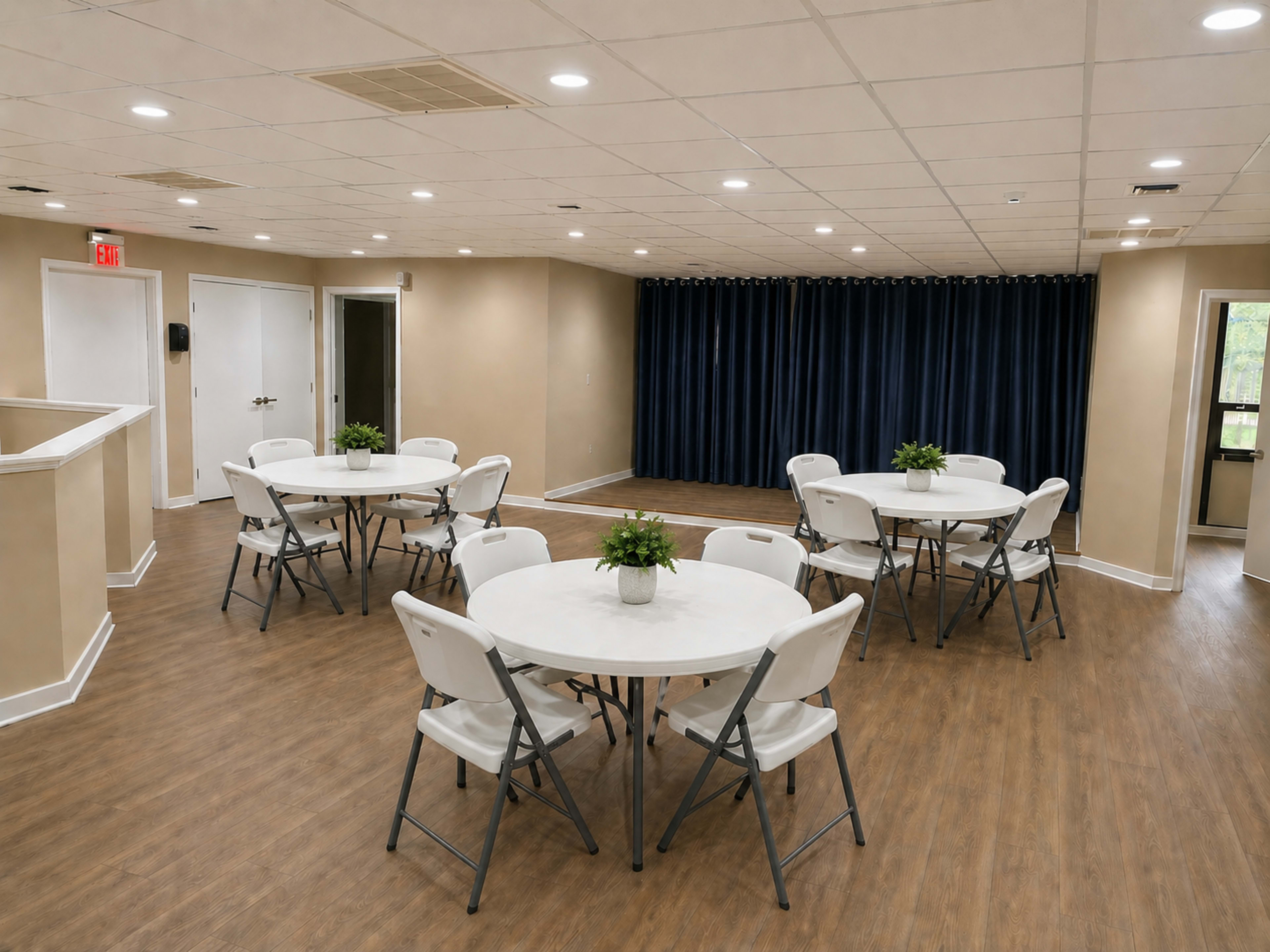 The image shows a spacious indoor room with several round tables and chairs arranged in a neat layout, featuring light-colored walls and a dark curtain at one end.