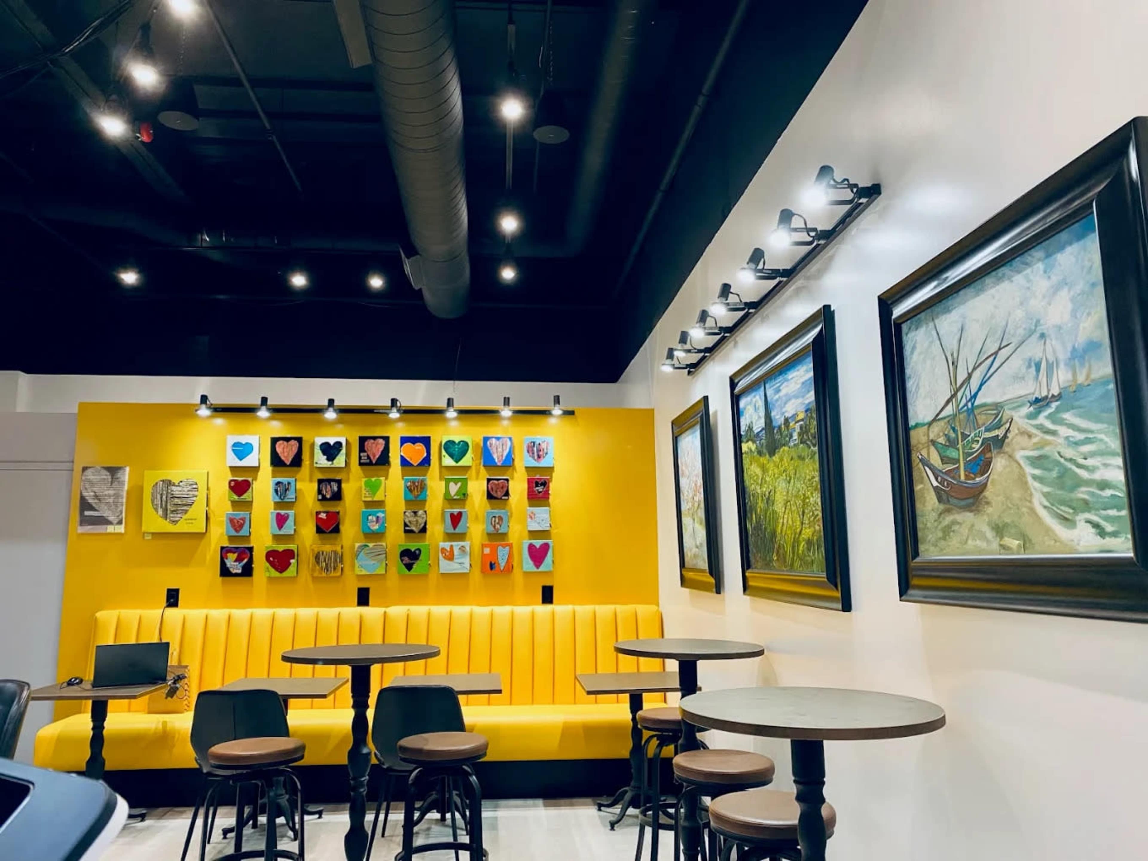 The image shows a modern cafe interior featuring yellow walls adorned with colorful artwork and a seating area with a yellow bench and circular tables.