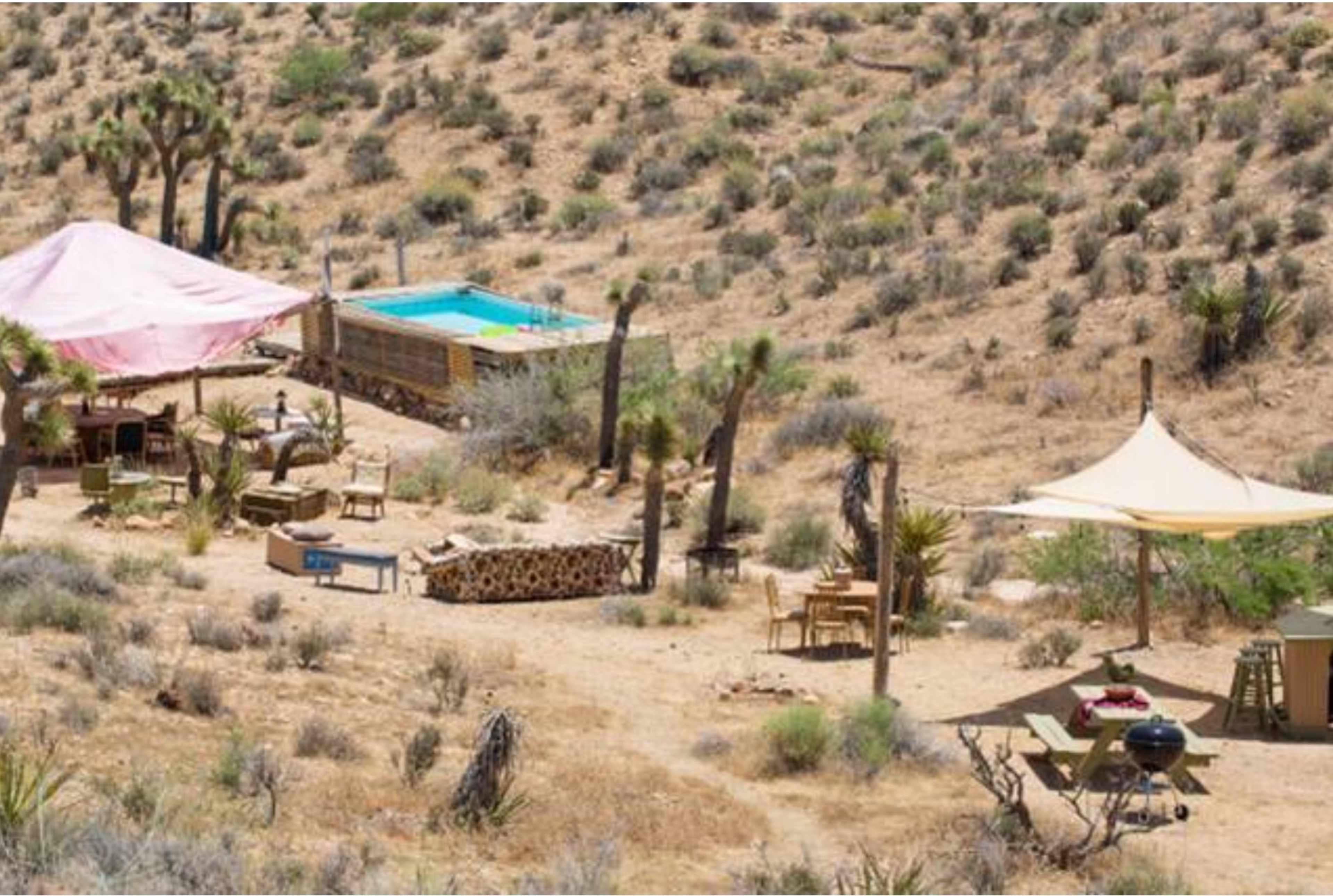 The image shows a desert landscape with a pink tent, a small swimming pool, and outdoor furniture scattered around.