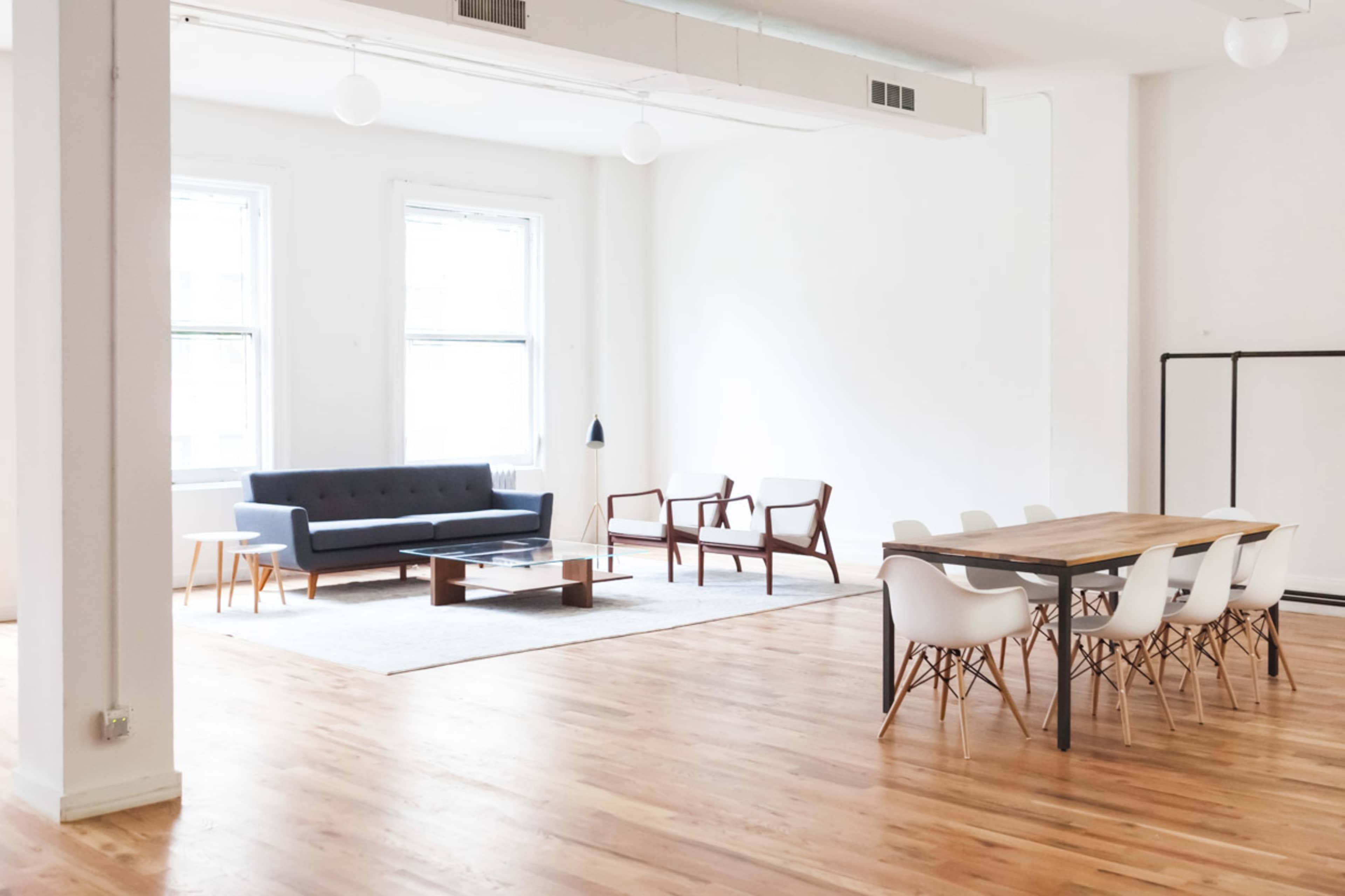 The image shows a spacious, minimalist interior featuring a seating area with a gray sofa and wooden chairs, alongside a dining table with white chairs.