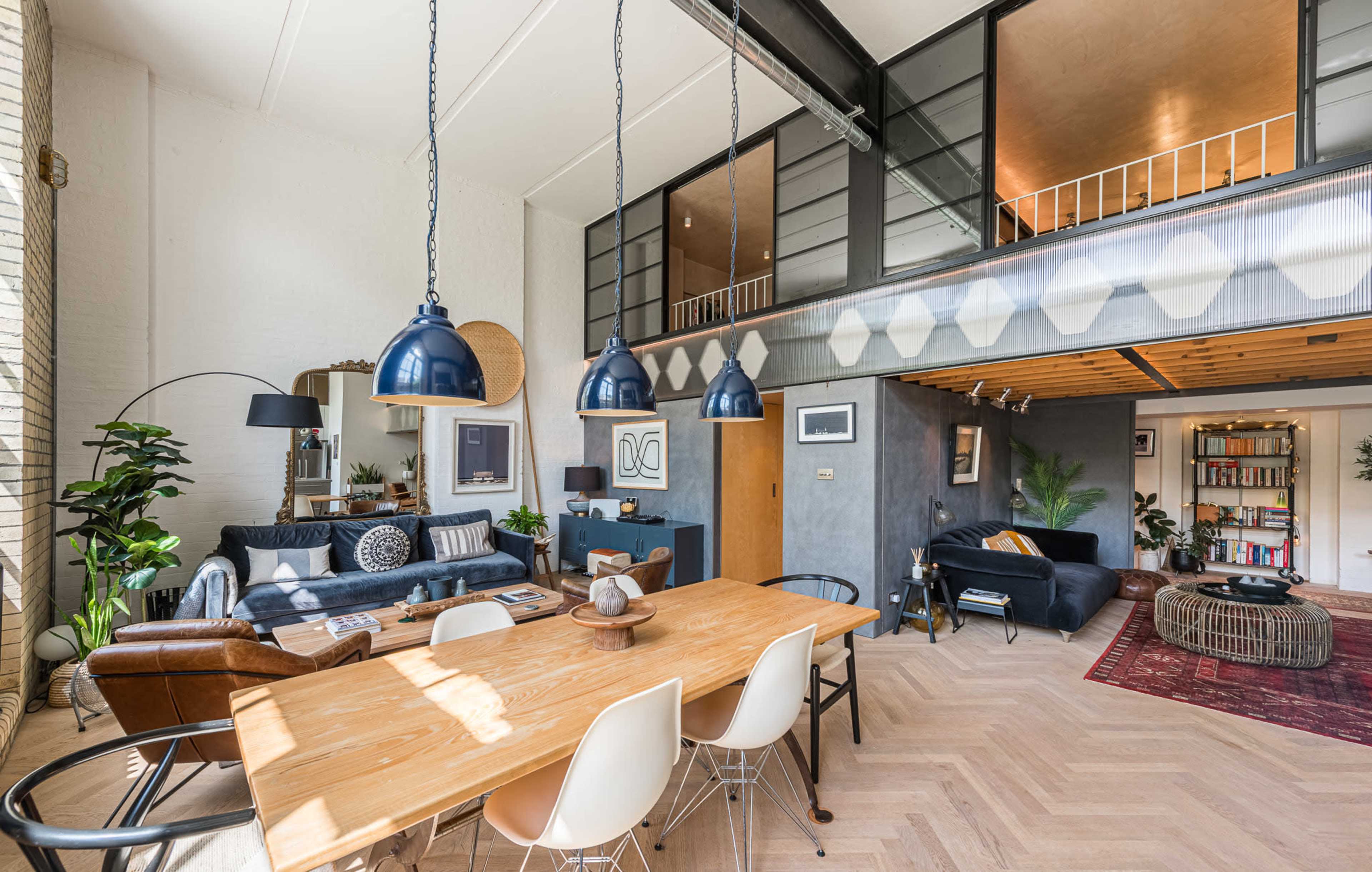 The image shows a spacious living area with a dining table, a leather couch, and various plants, featuring modern decor and a lofted section above.