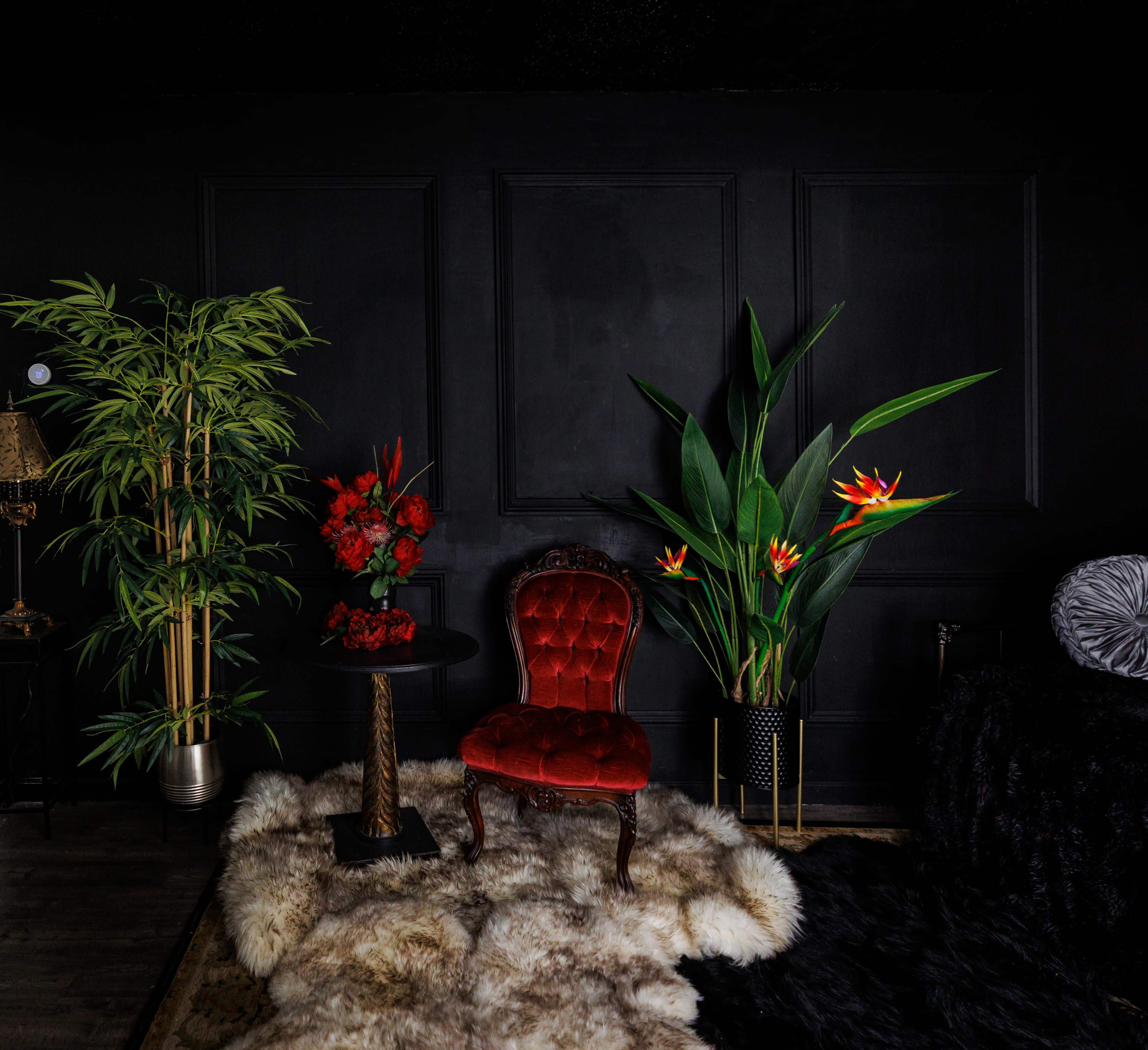 The image shows a cozy interior featuring a red velvet chair, a black side table with flowers, and tall green plants against a dark wall.