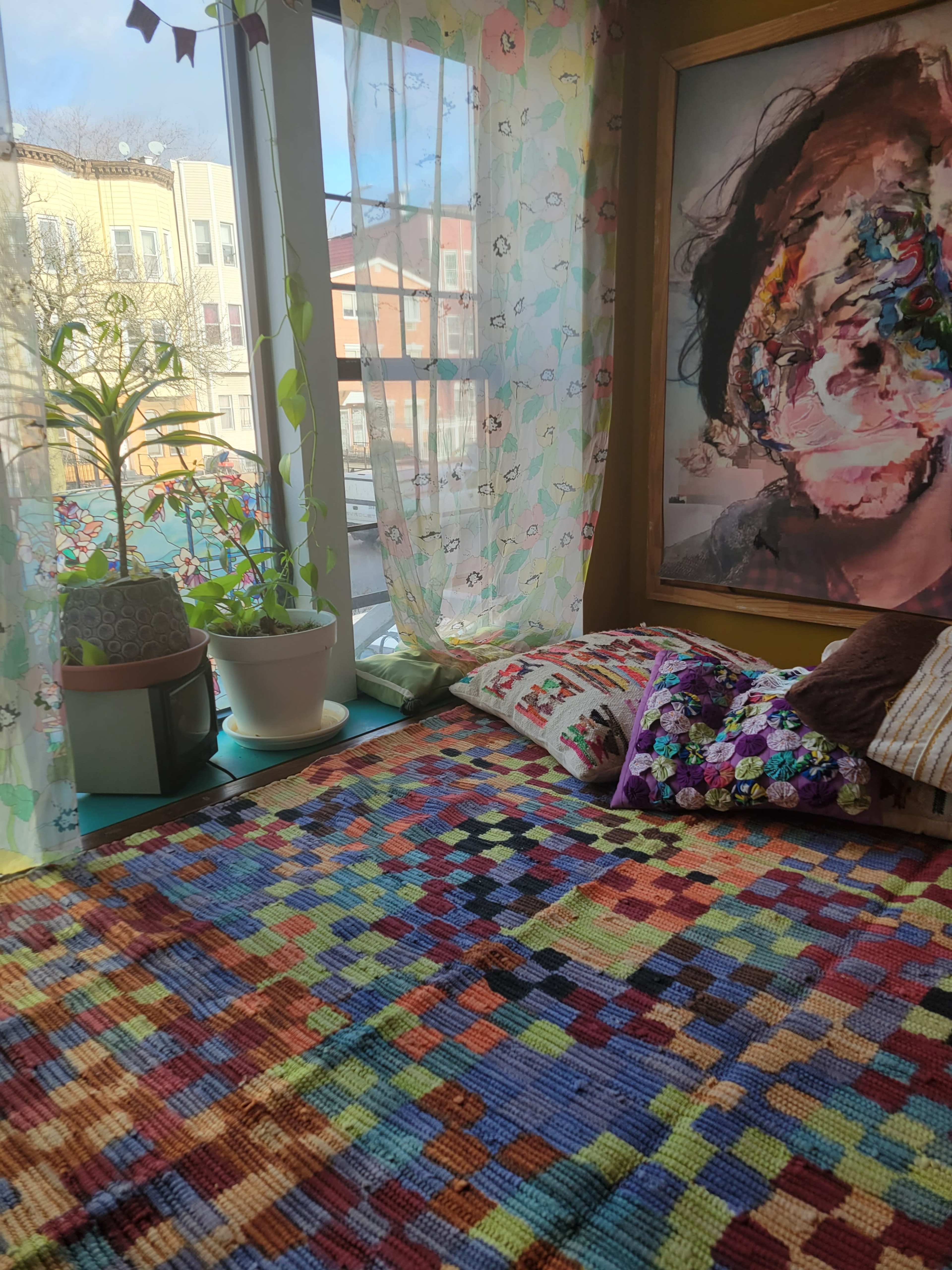 A colorful, patterned rug covers the floor in a sunlit room with a large window, plants, and a framed portrait on the wall.