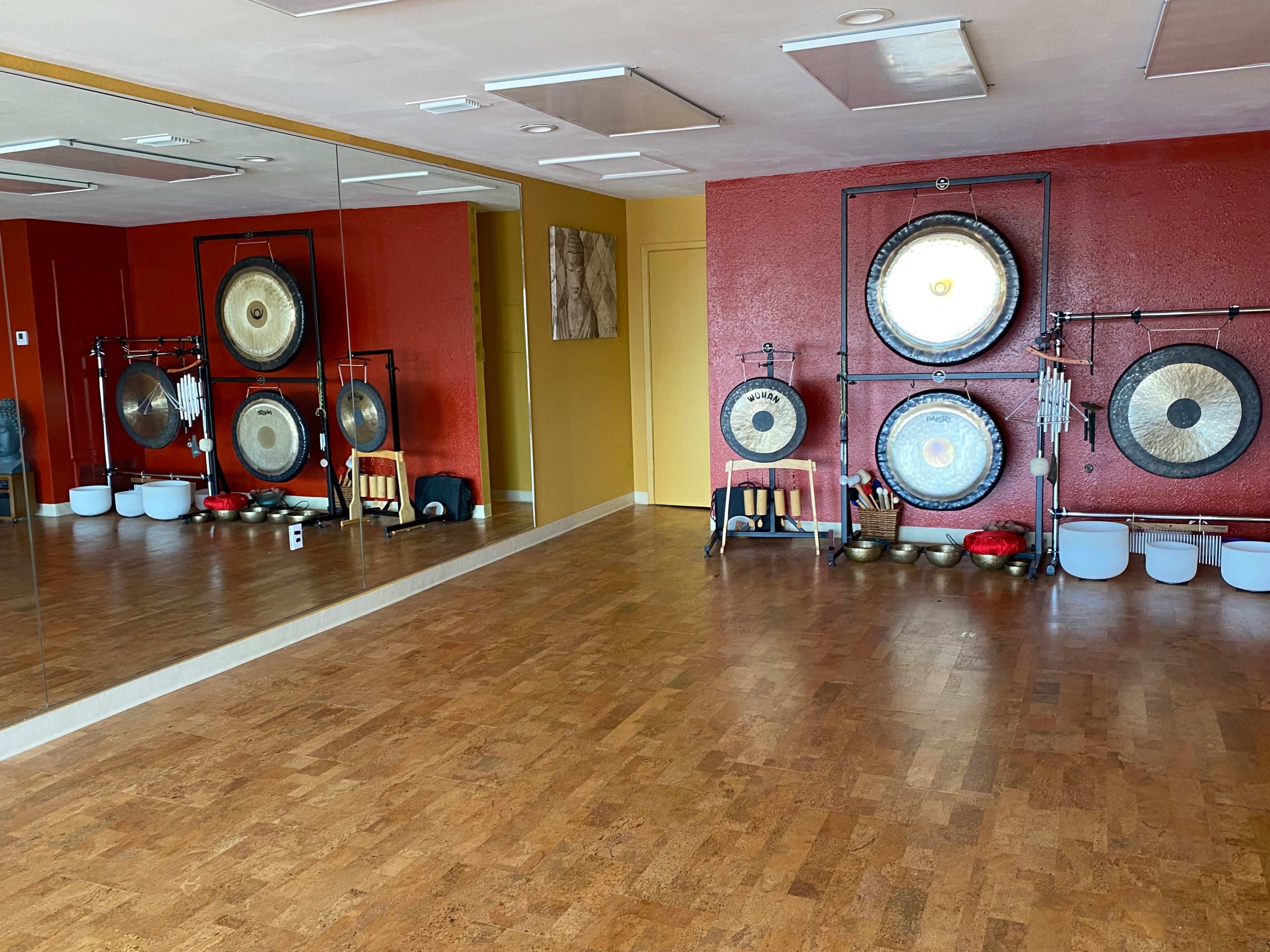 The image shows a spacious room with wooden floors and red walls, featuring a set of gongs and bowls arranged against a mirrored wall.