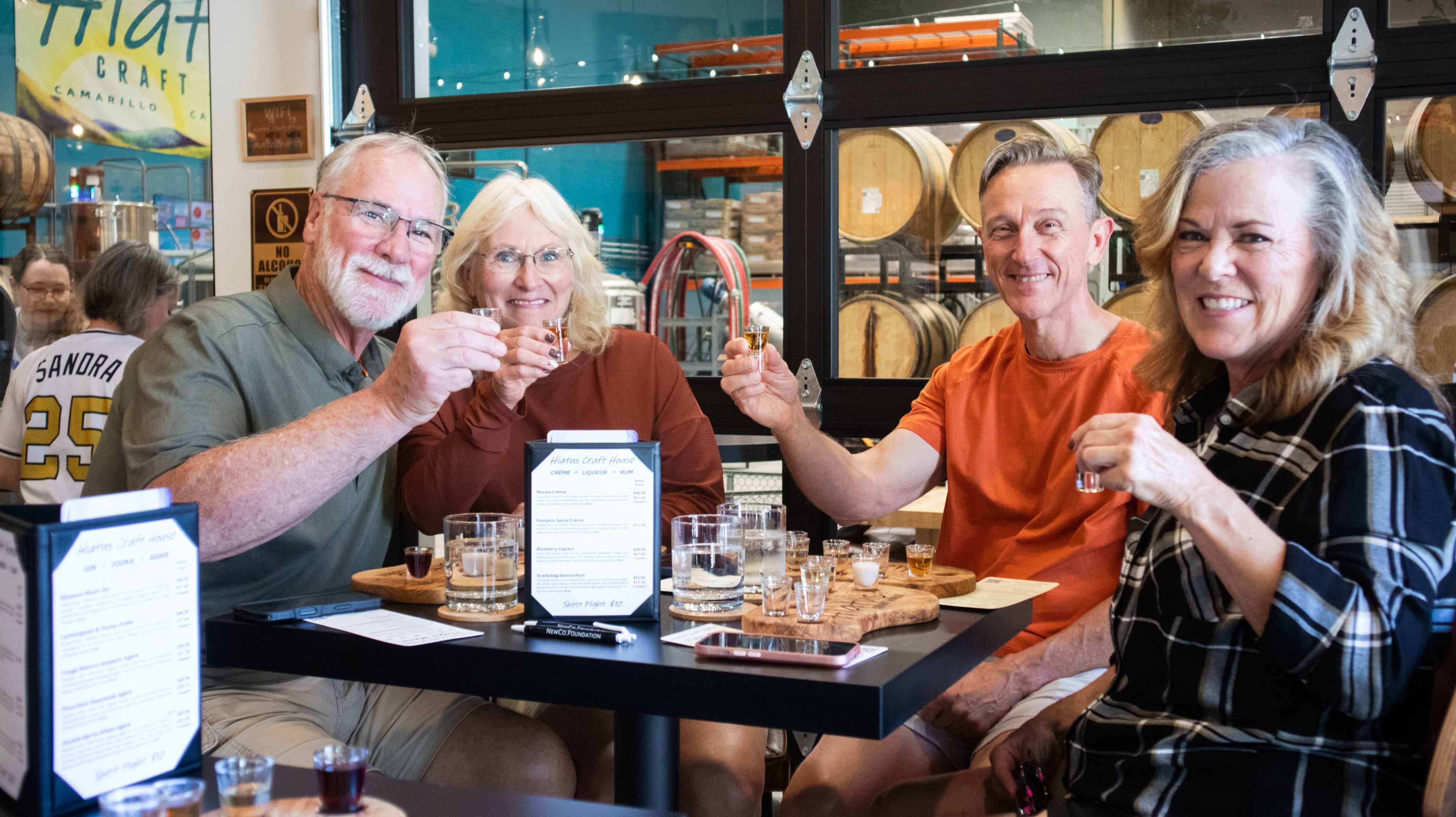 Four people are gathered around a table, tasting samples of drinks in a brewery with wooden barrels in the background.