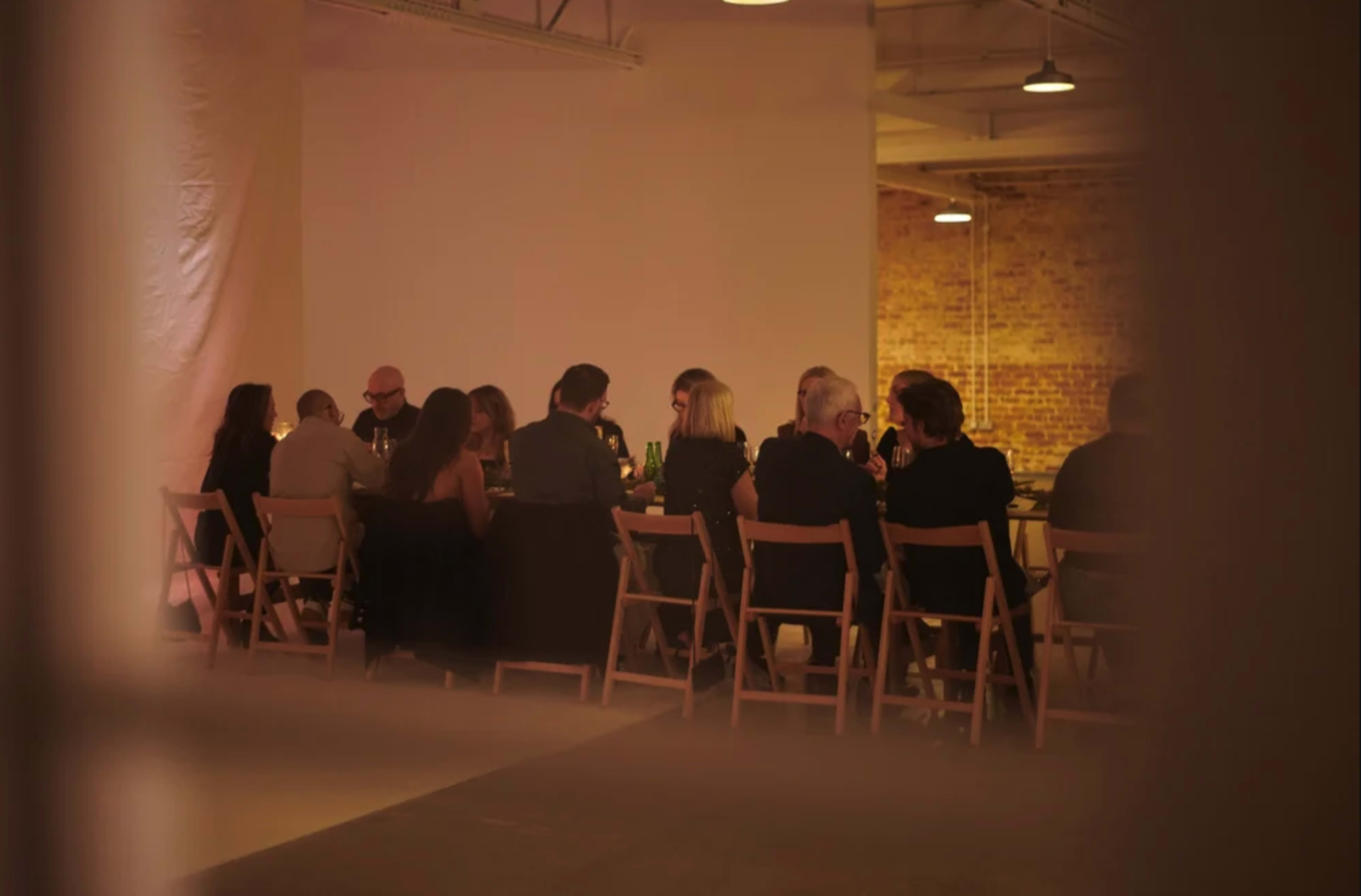 A group of people is seated around a long table in a dimly lit room, sharing a meal or conversation.