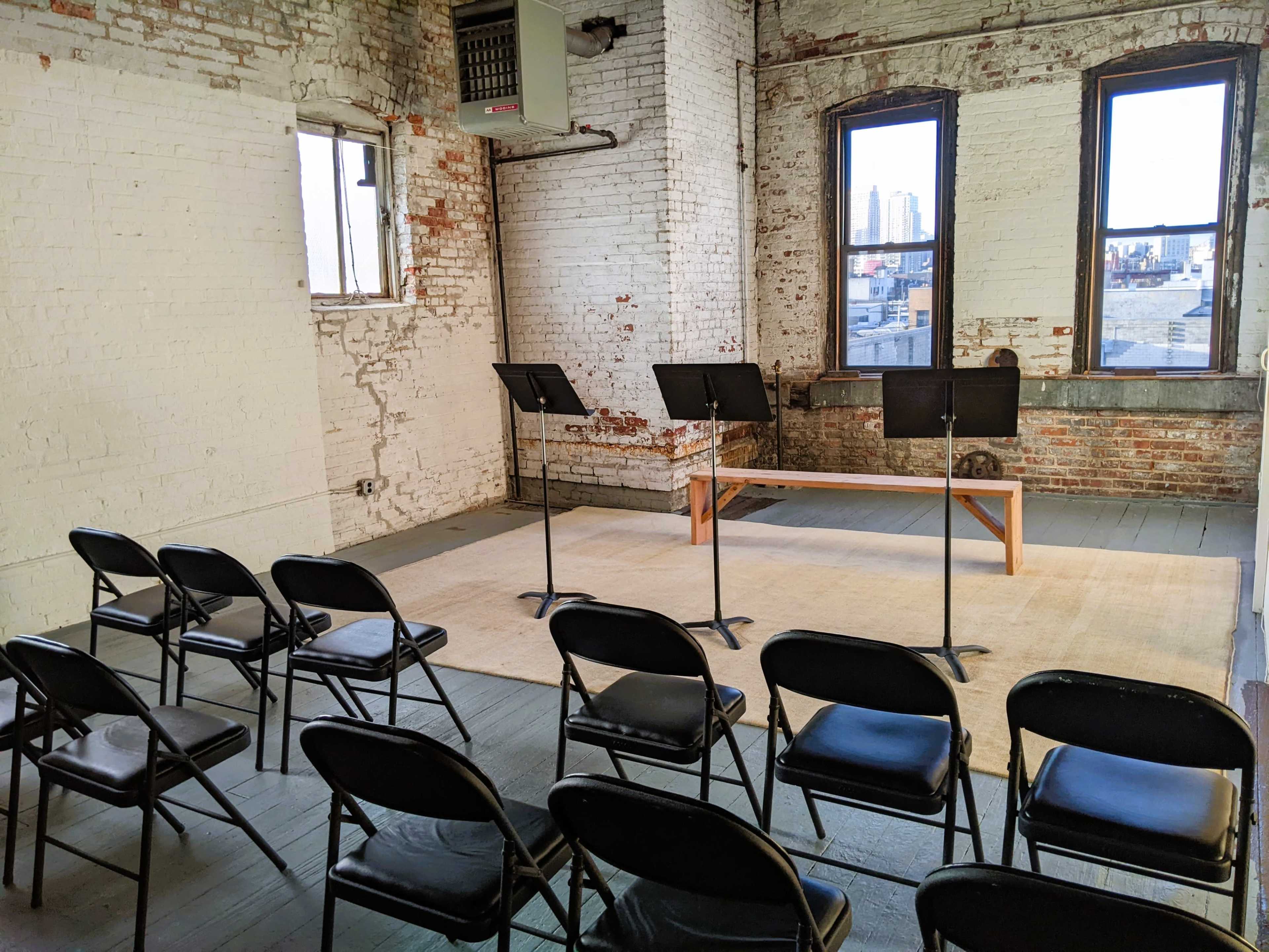 A small, empty performance space features a wooden platform and several music stands arranged in front of rows of black chairs.