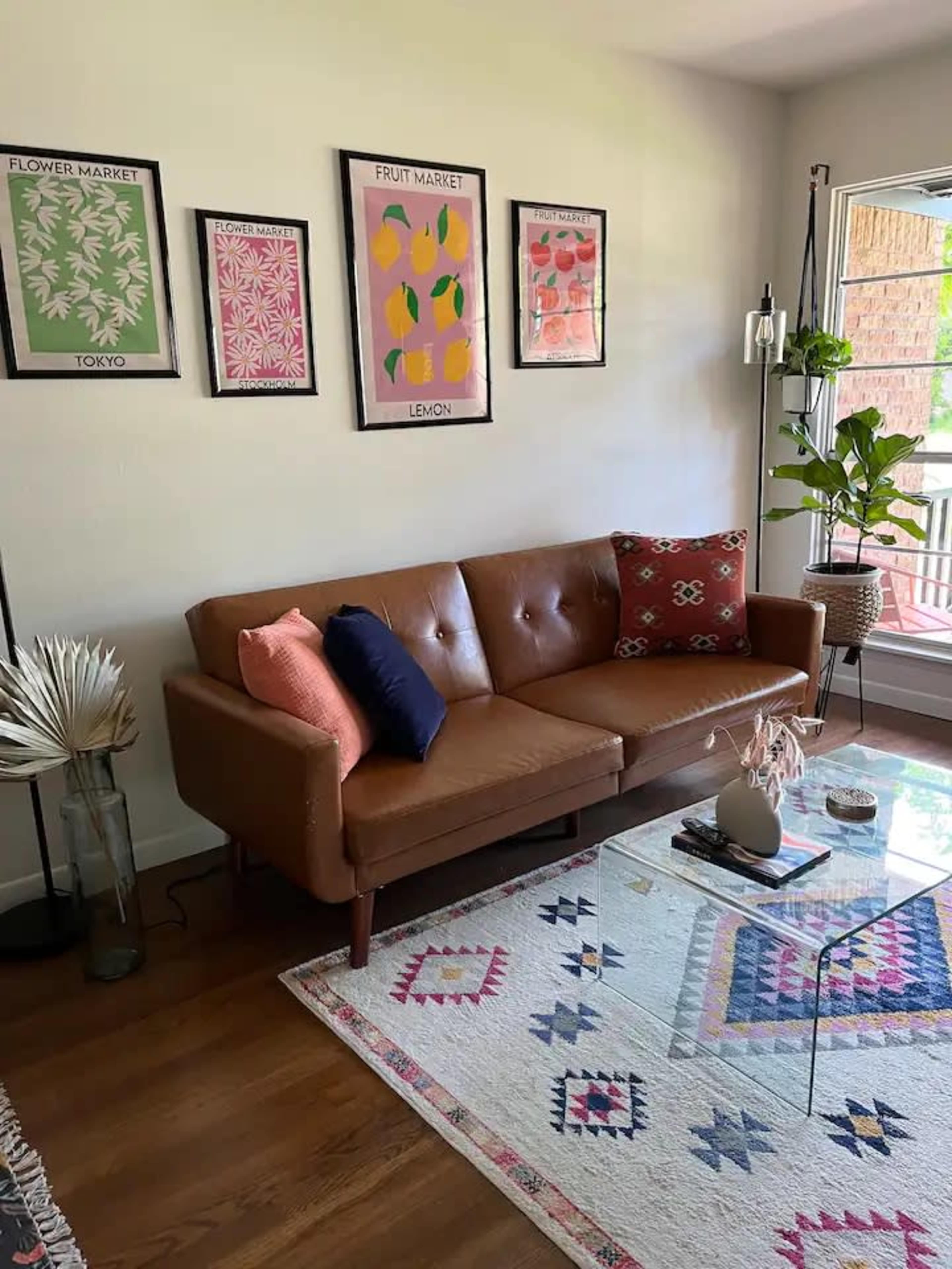 A brown leather sofa with colorful cushions positioned near a window, complemented by framed botanical prints on the wall and a glass coffee table.