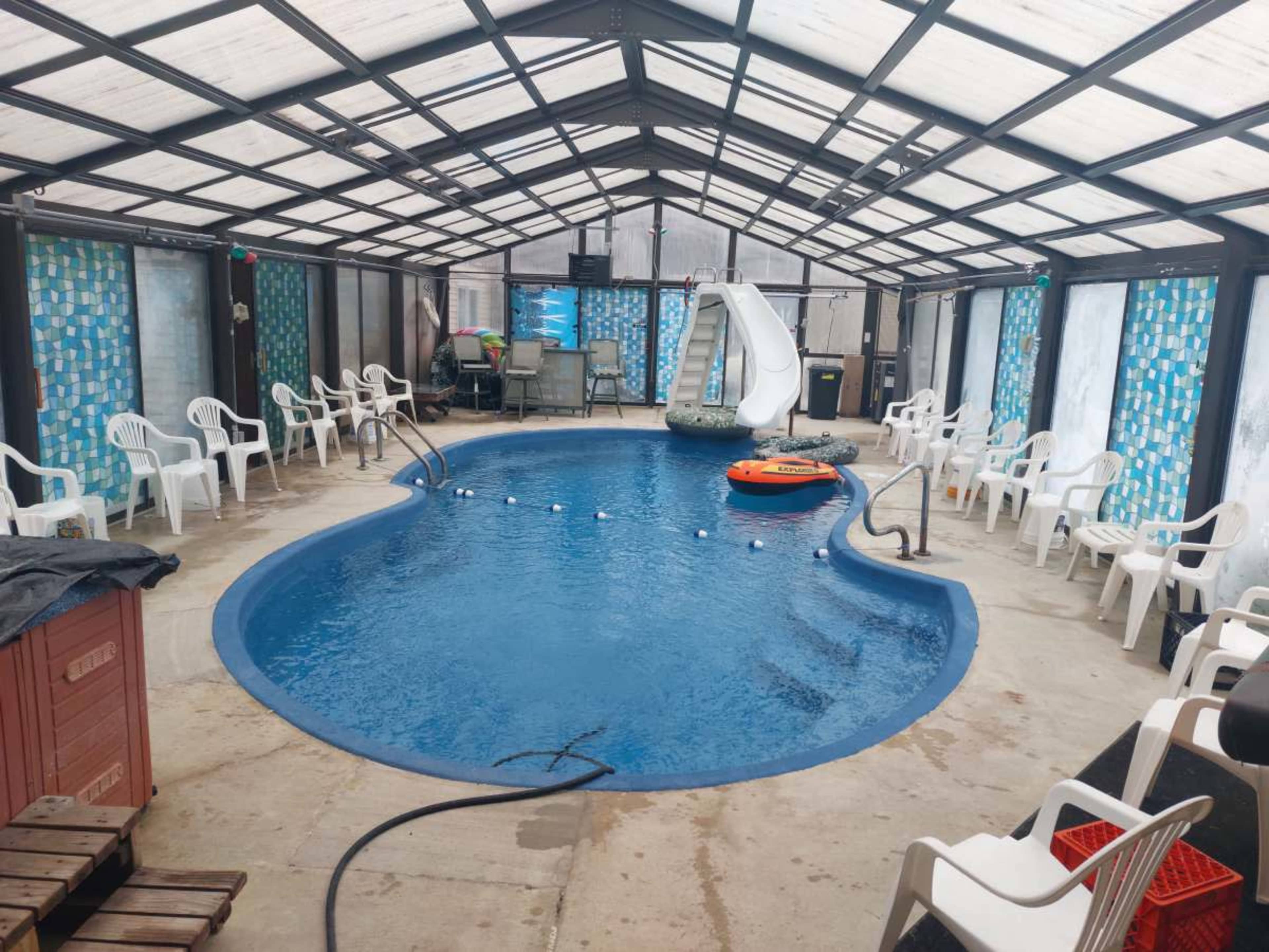 An indoor swimming pool enclosed by a glass roof, surrounded by white plastic chairs and a small play slide.