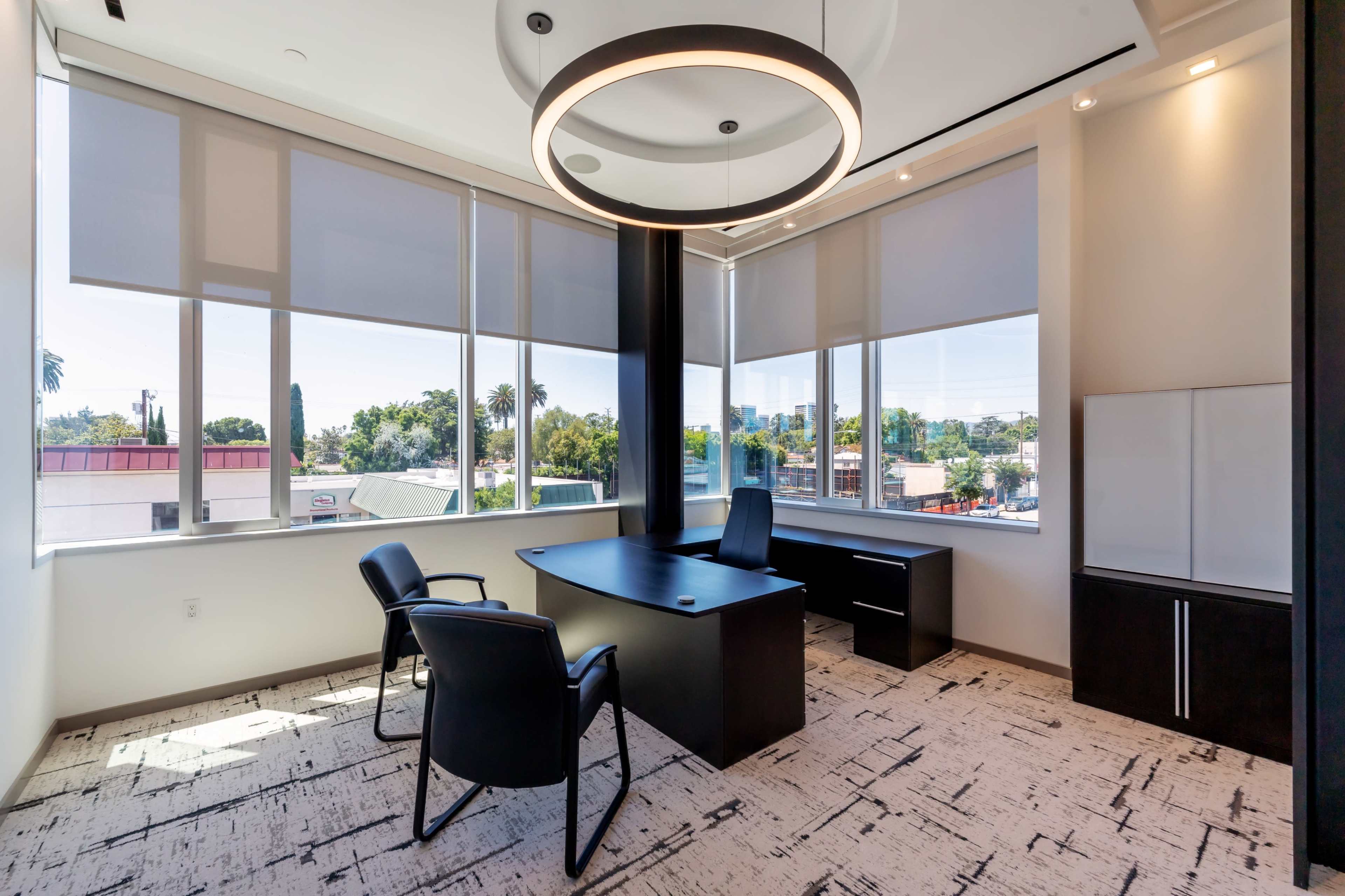 A modern office space features a black desk and two chairs, with large windows allowing natural light and a view of the cityscape.