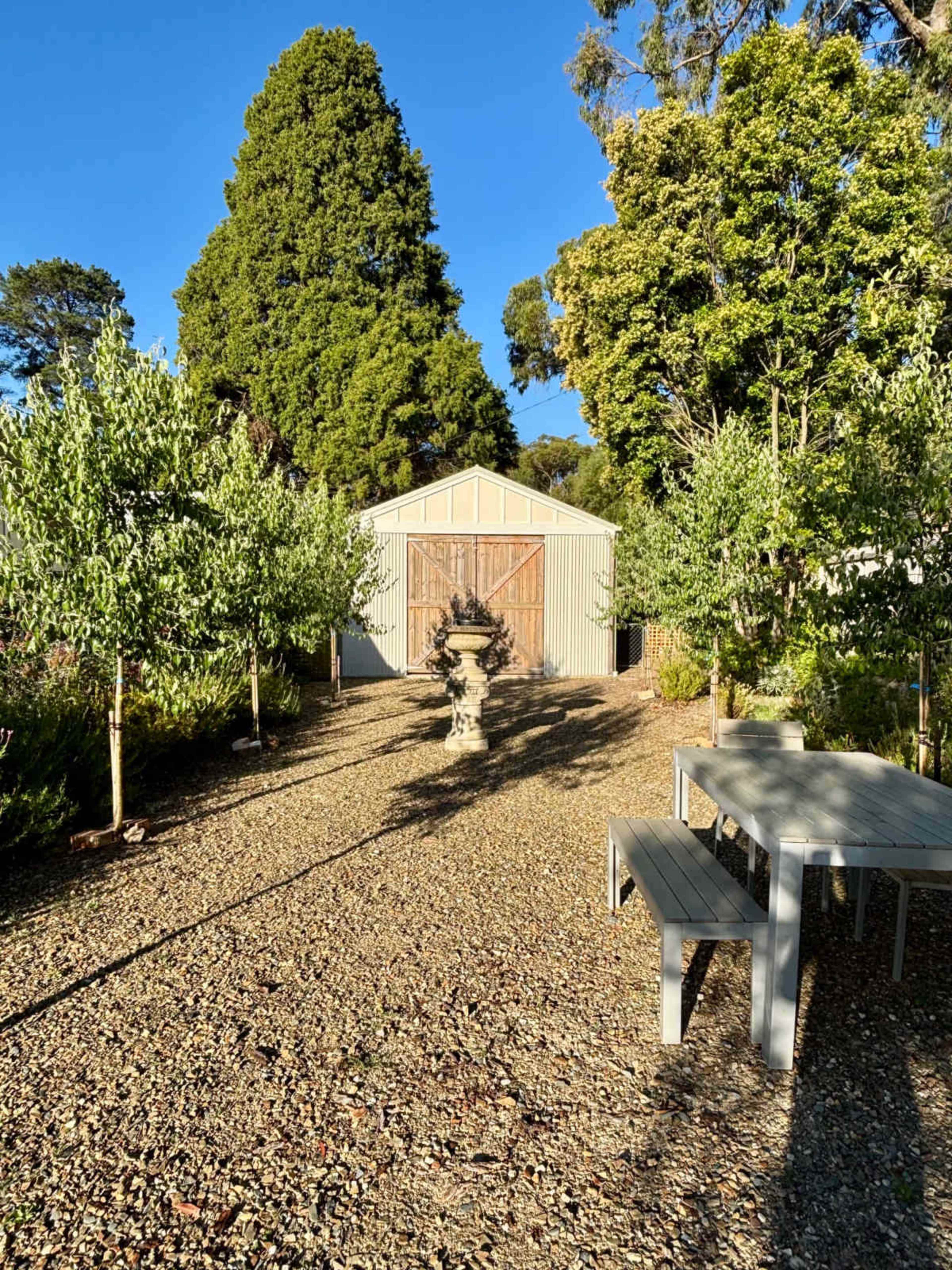 A gravel pathway leads to a garden shed surrounded by trees and a stone fountain, with a gray outdoor table positioned nearby.