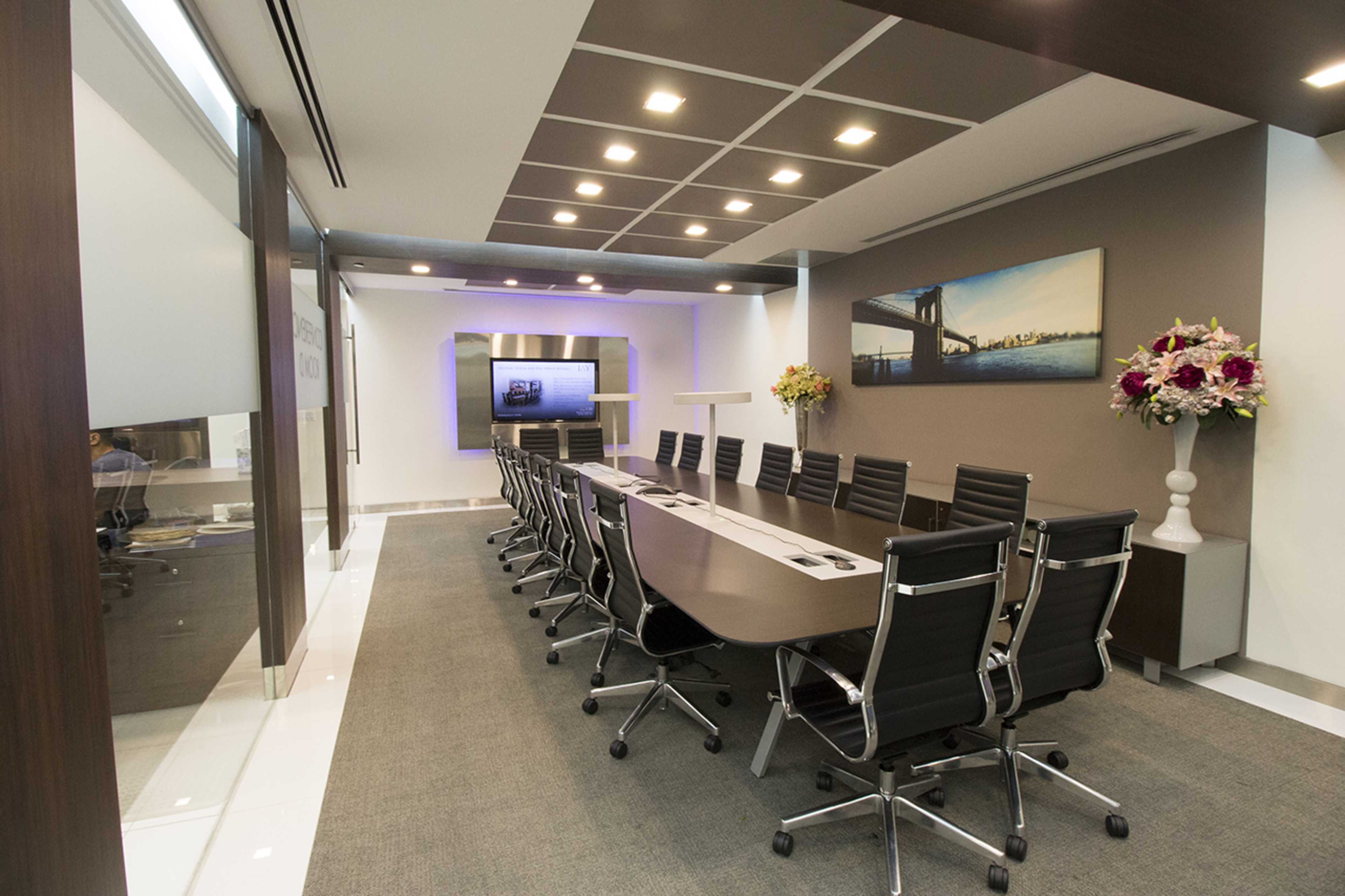 A modern conference room with a long table, black chairs, a wall-mounted television, and decorative flowers.