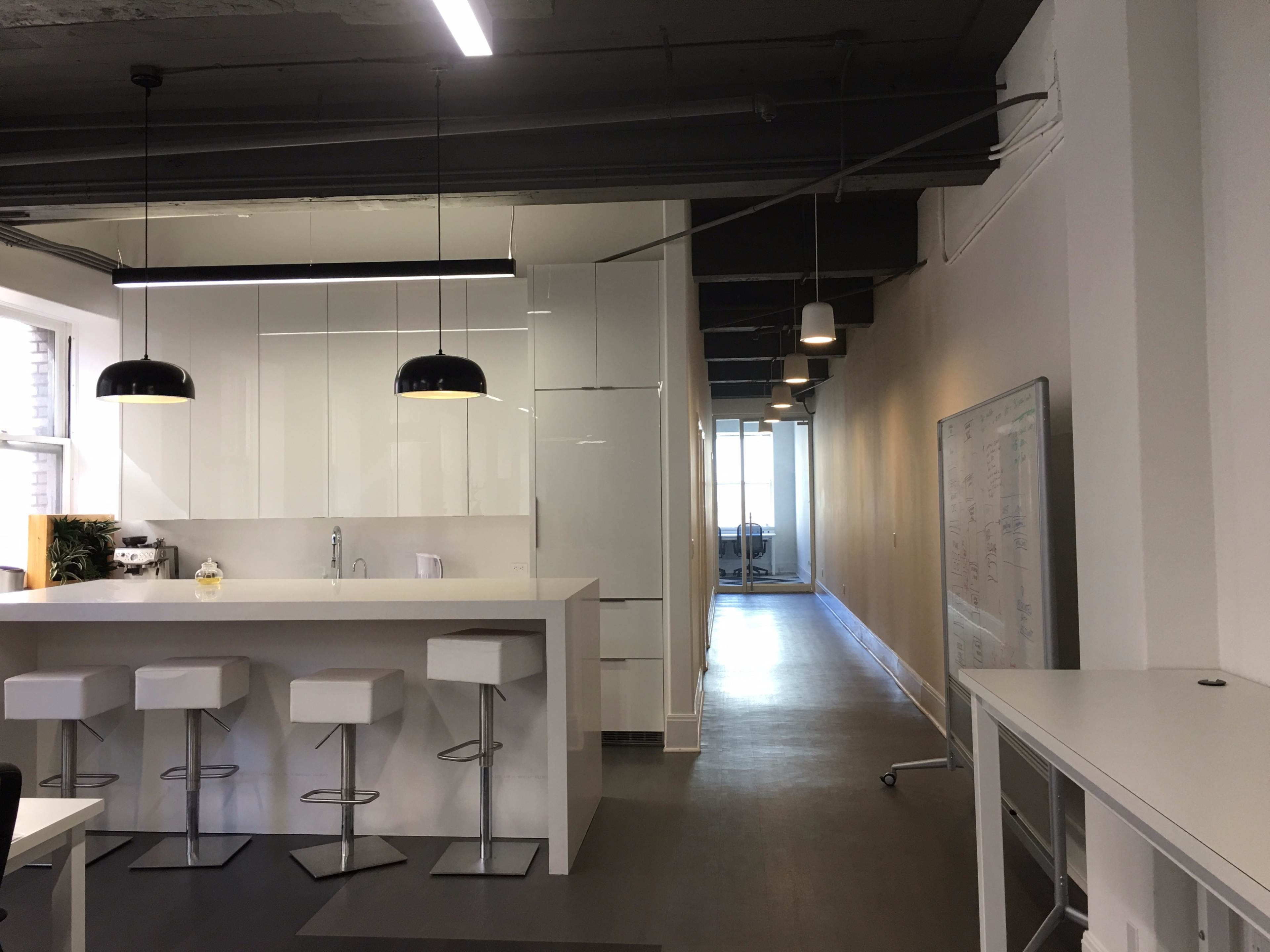 A modern office space featuring a white kitchen island with bar stools, bright lighting, and a corridor leading to a large window at the far end.