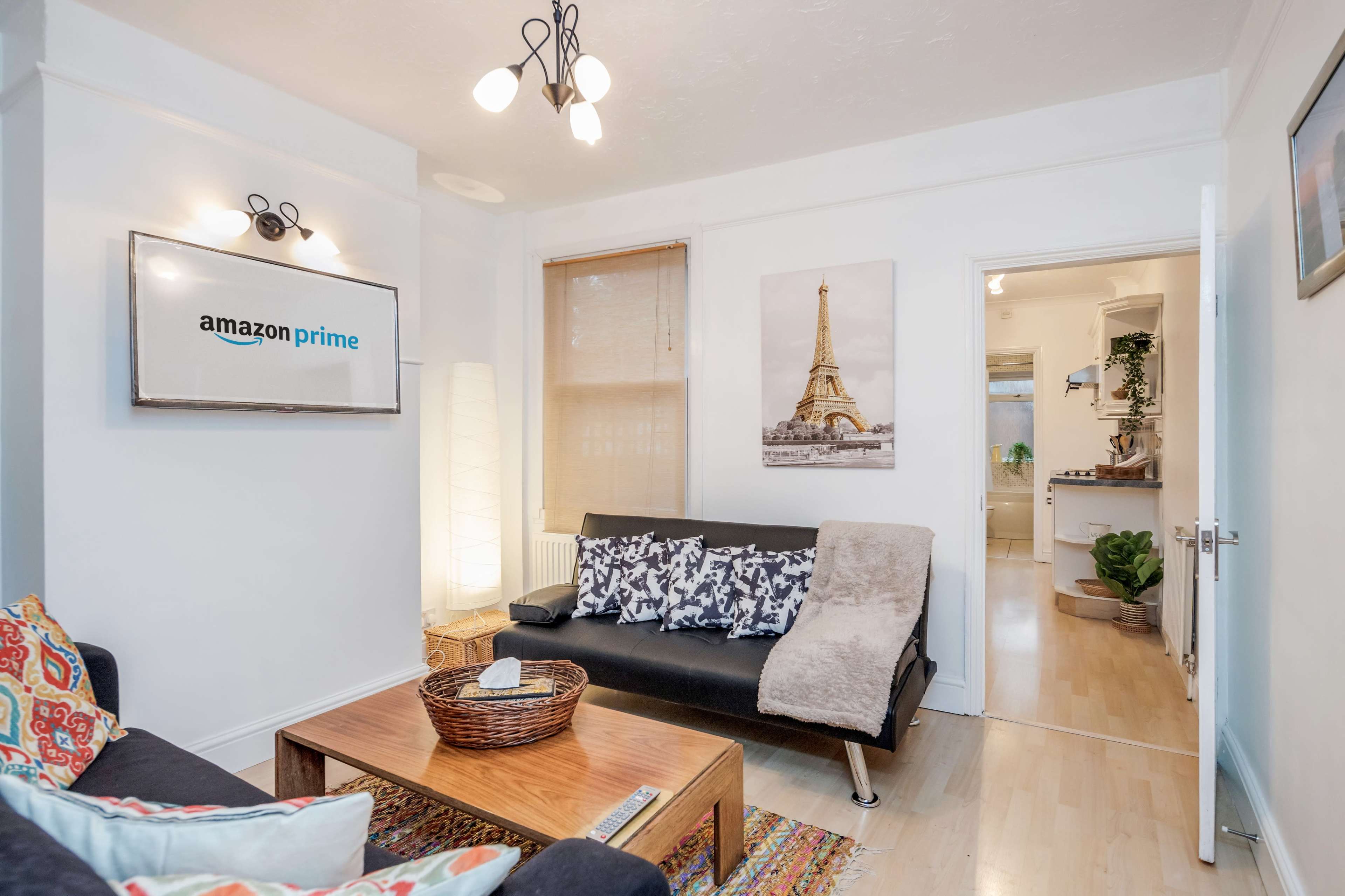 A living room with a black couch, a coffee table, an Amazon Prime sign on the wall, and a doorway leading to a kitchen area.