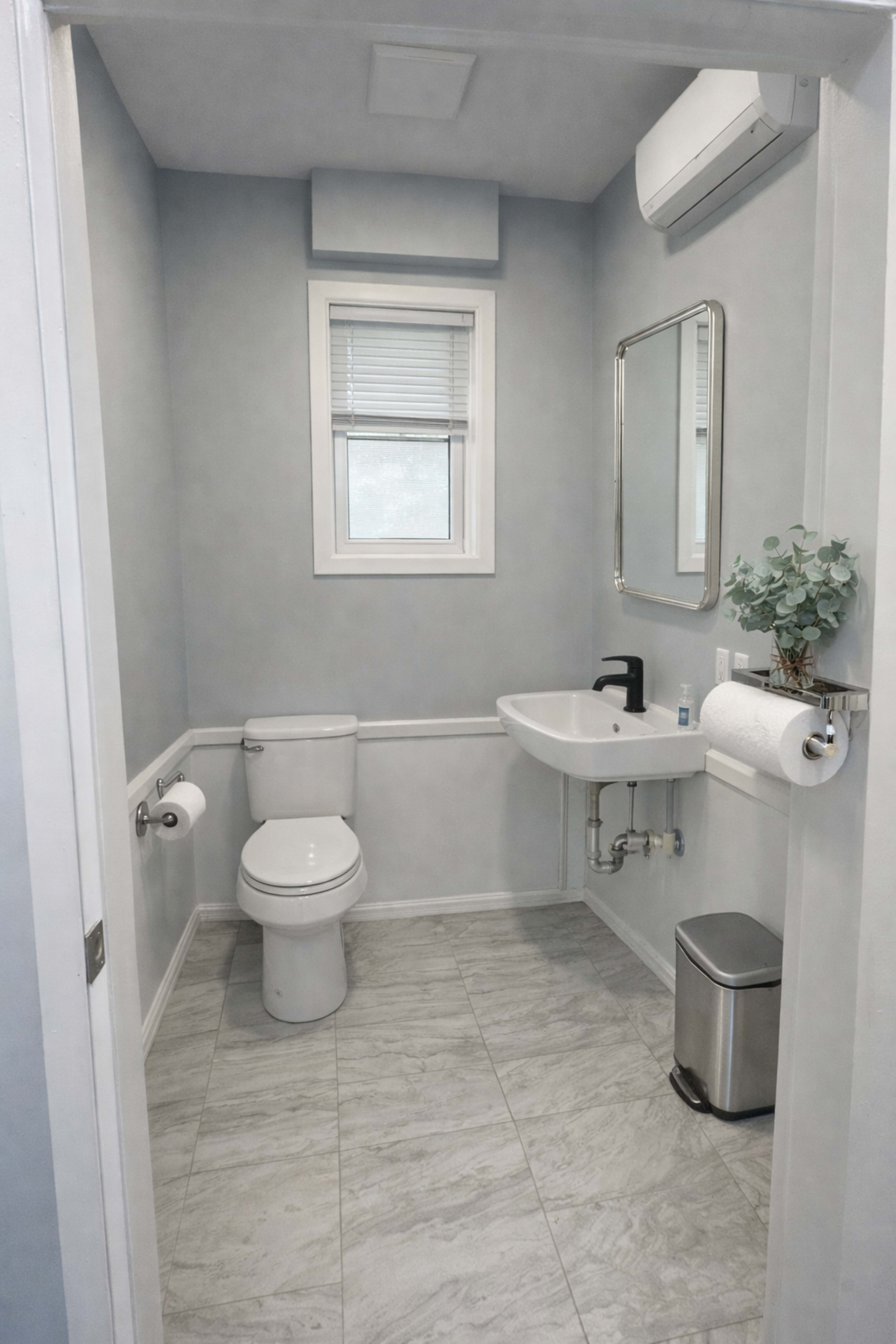 The image shows a small, modern bathroom featuring a toilet, a wall-mounted sink, a mirror, and a small window.