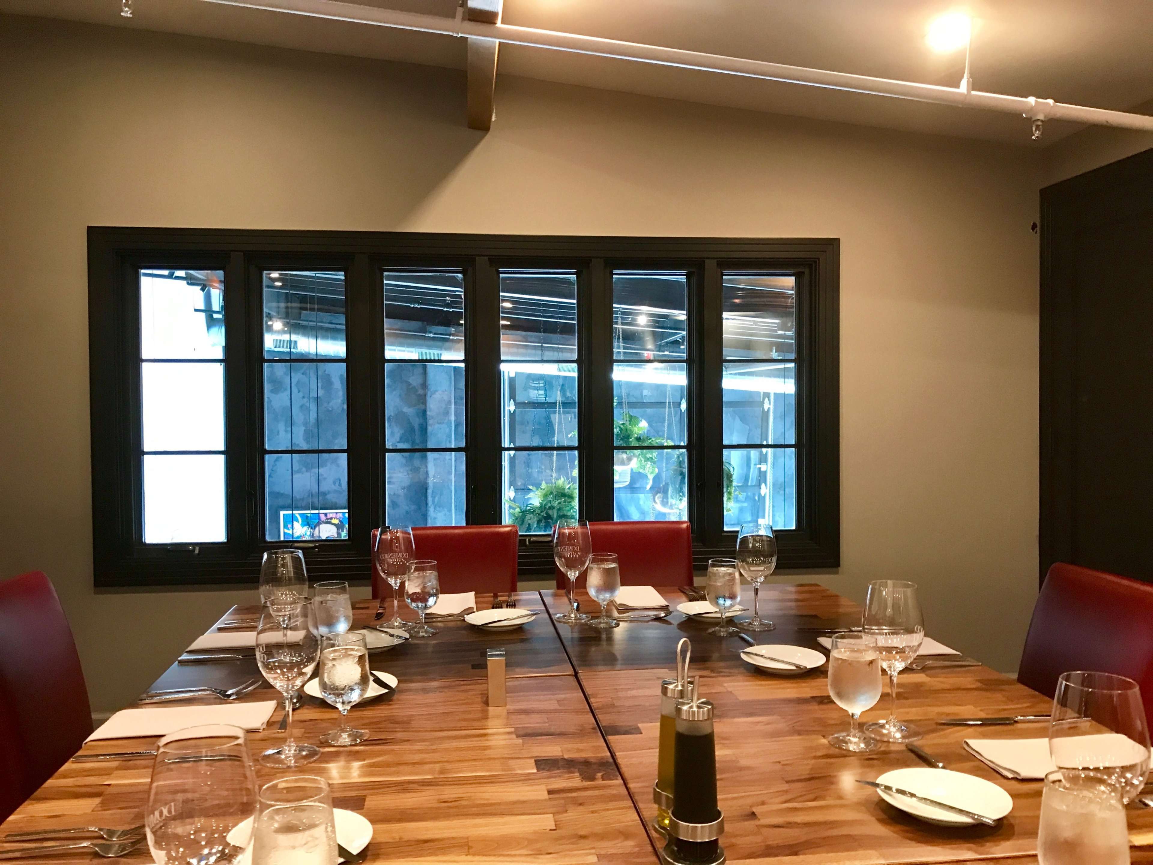A dining table is set with glassware and plates in a room with large windows showing a view outside.