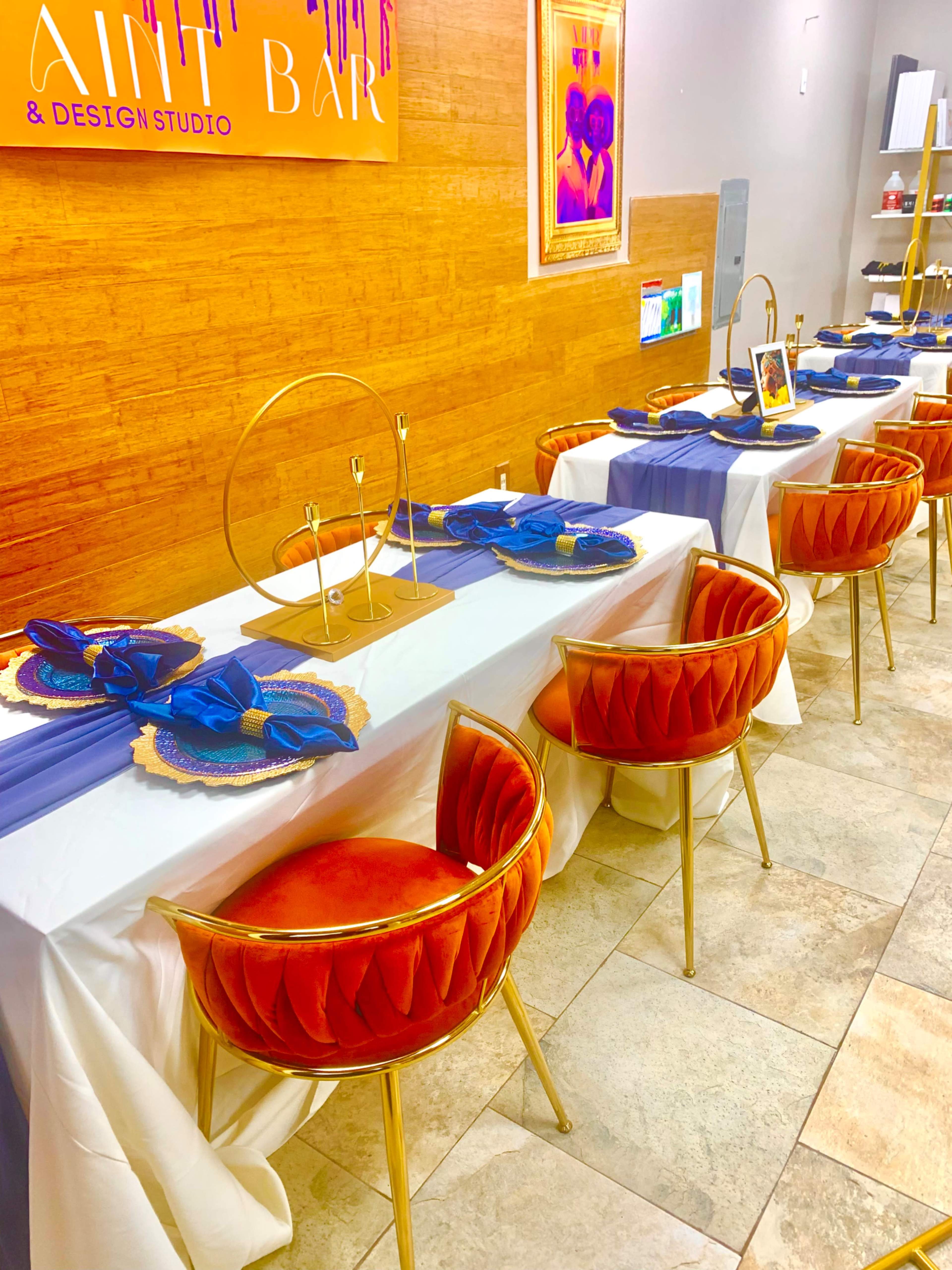 A dining setup with a long table covered in white and purple tablecloths, adorned with gold and blue tableware and velvet chairs.