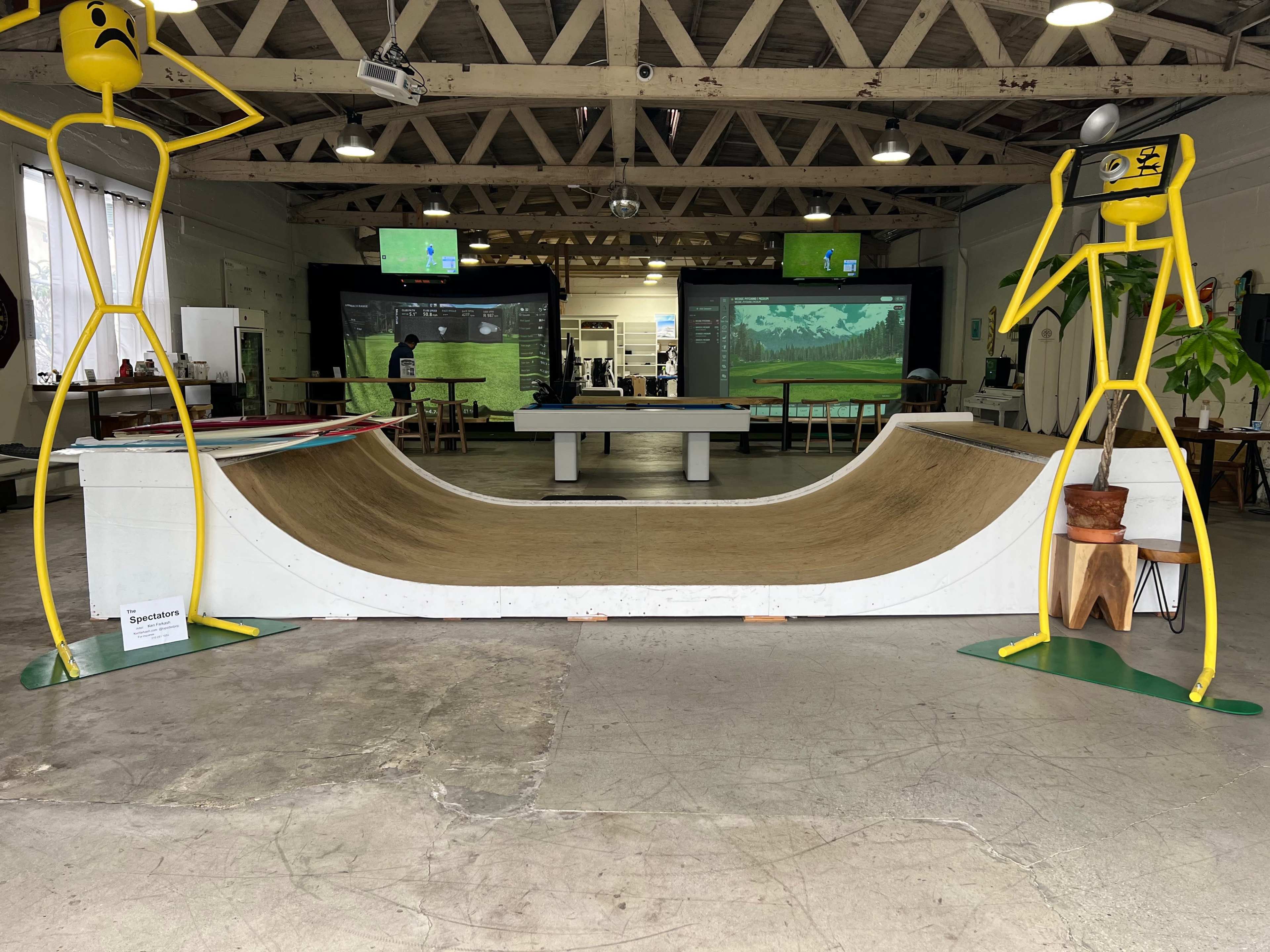 The image features a skateboard half-pipe ramp positioned in a spacious indoor venue, flanked by yellow, stick-figure sculptures.