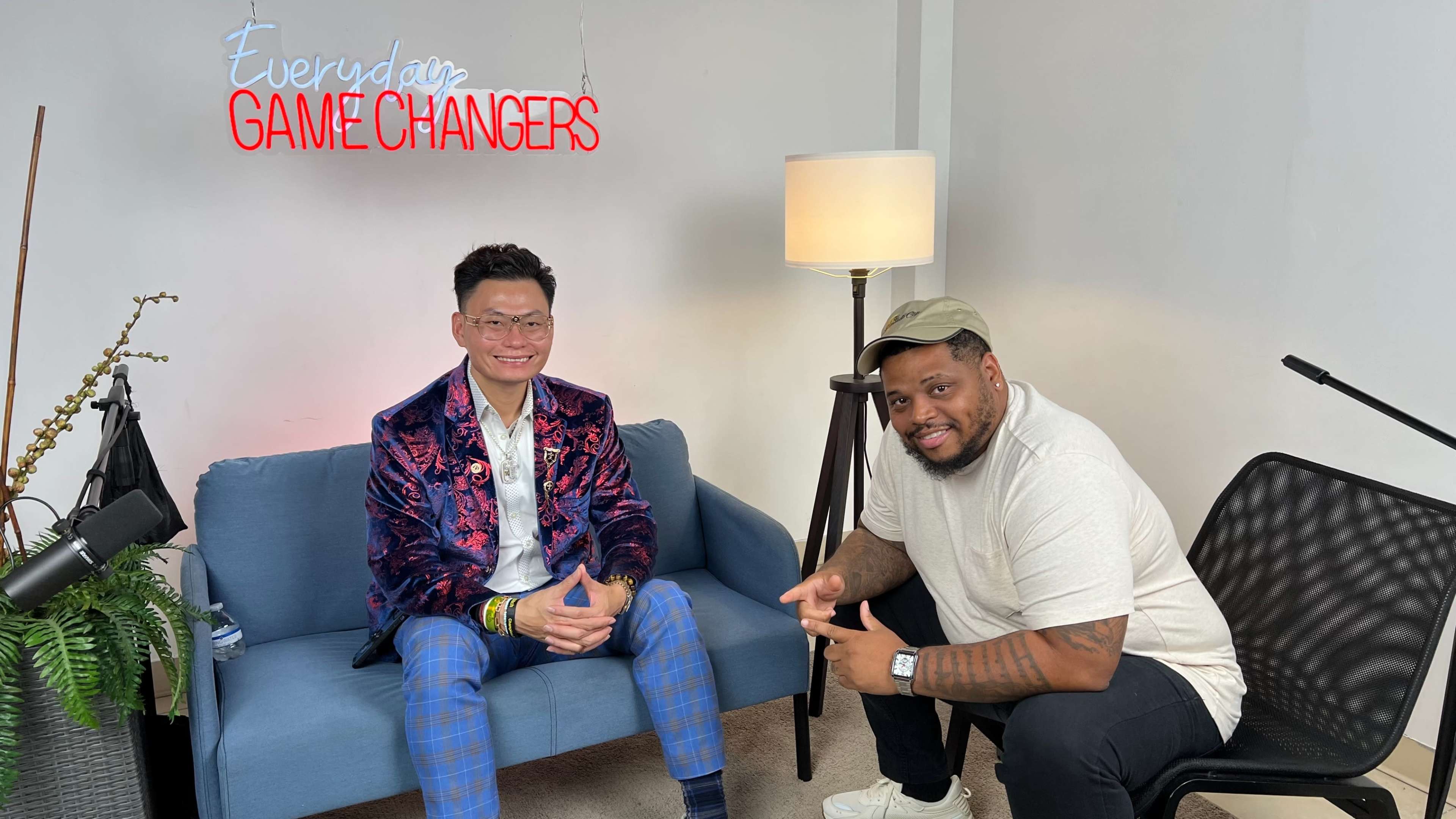 Two men are seated in a brightly lit studio, with one man in a colorful suit and the other in a casual outfit, posing for the camera in front of a decorative neon sign that reads "Everyday Game Changers."