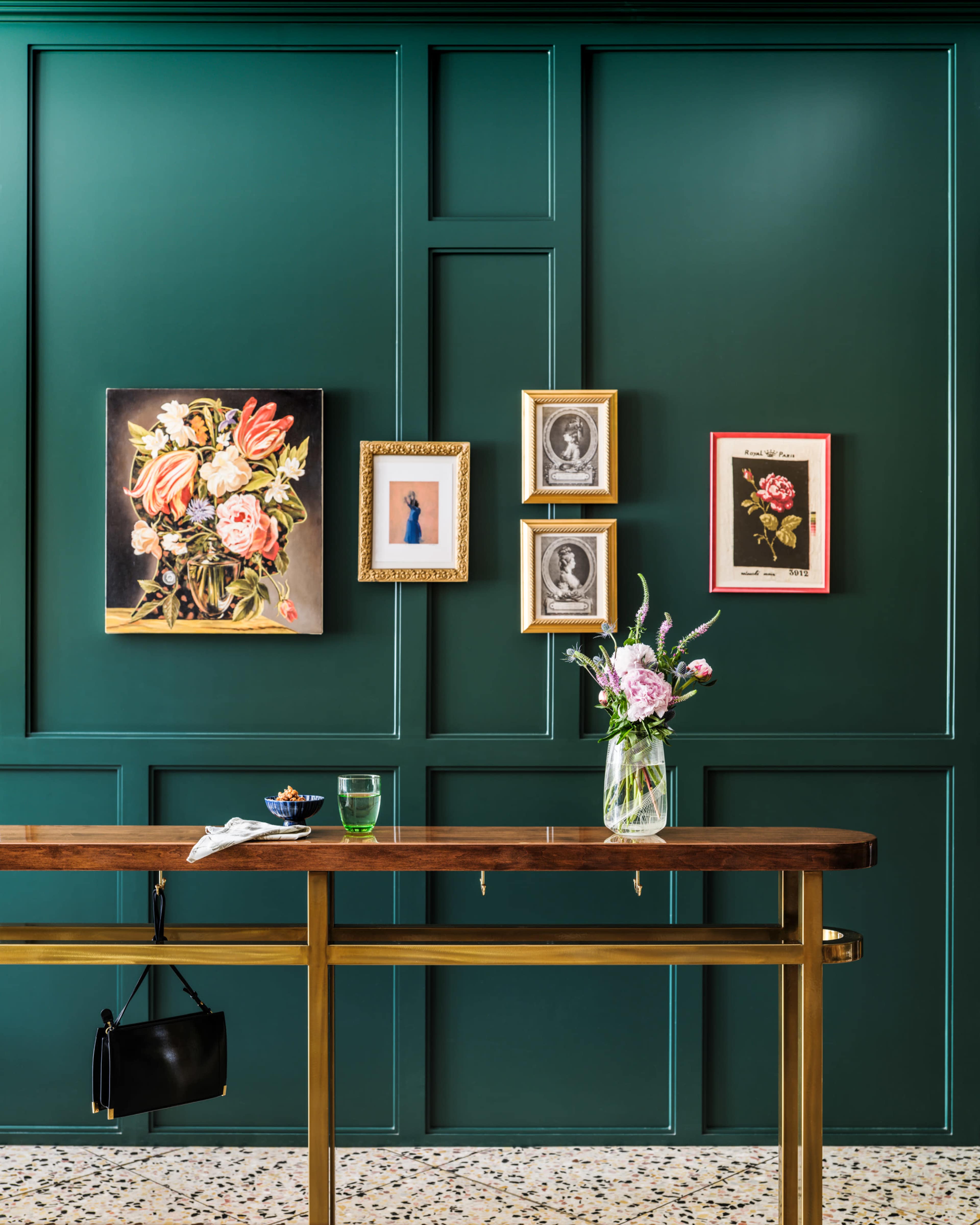 The image shows a stylish interior featuring a dark green wall adorned with various framed artworks, alongside a wooden table with a vase of flowers and a black handbag hanging from it.