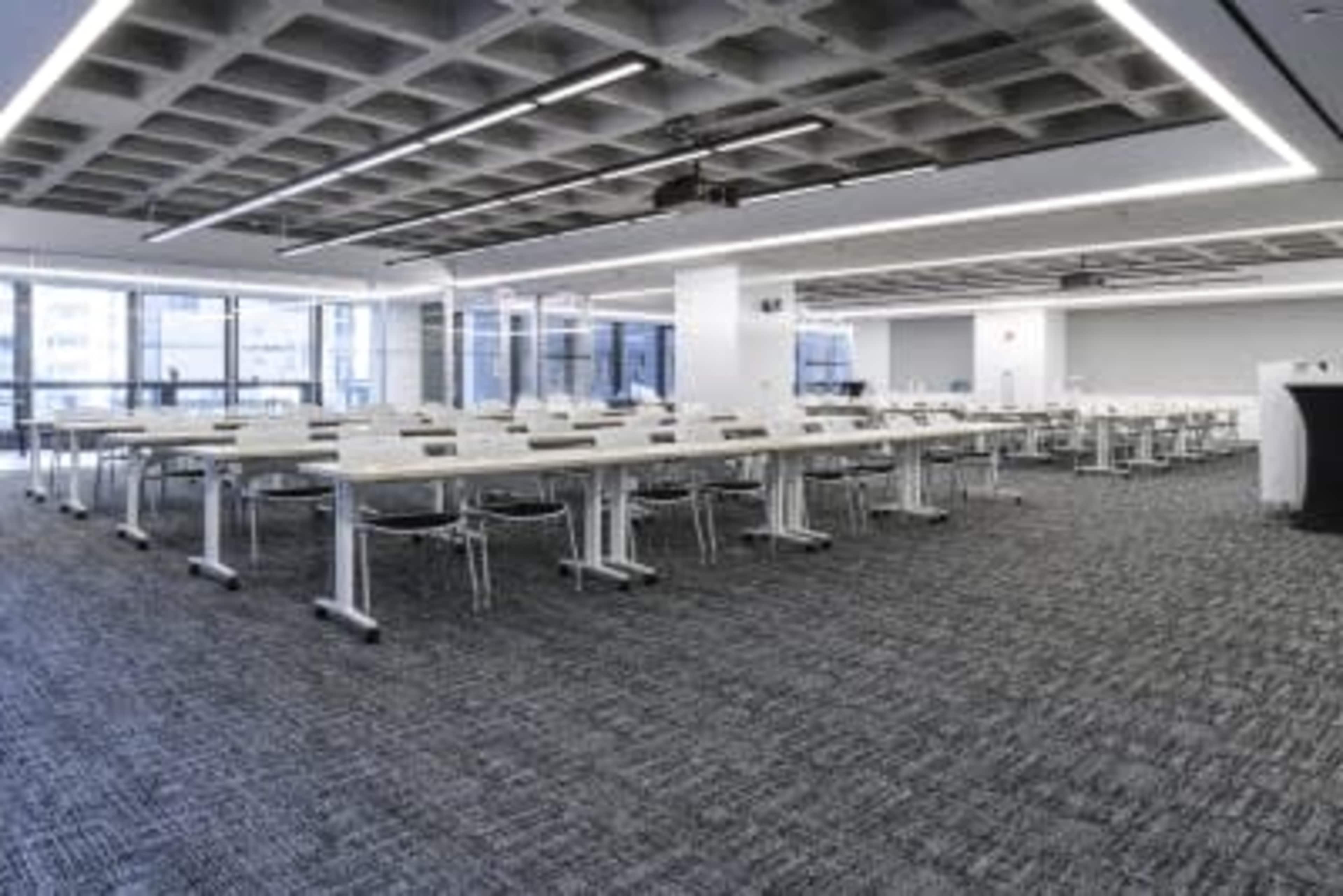The image shows a modern conference room with rows of tables and chairs arranged for an event, featuring large windows that provide natural light.