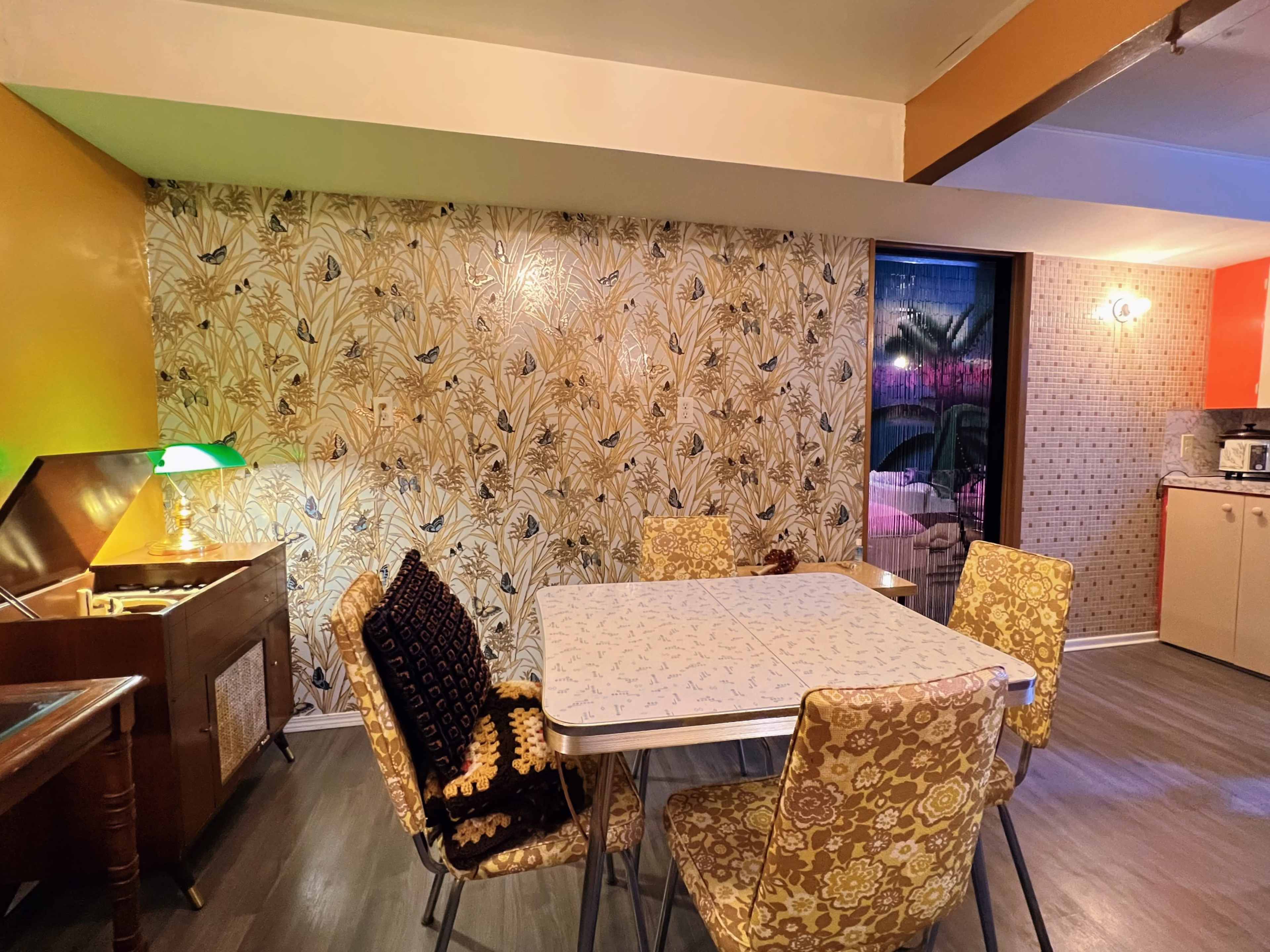 A dining area with a patterned wallpaper, a table surrounded by six chairs, and a sideboard with a green lamp.