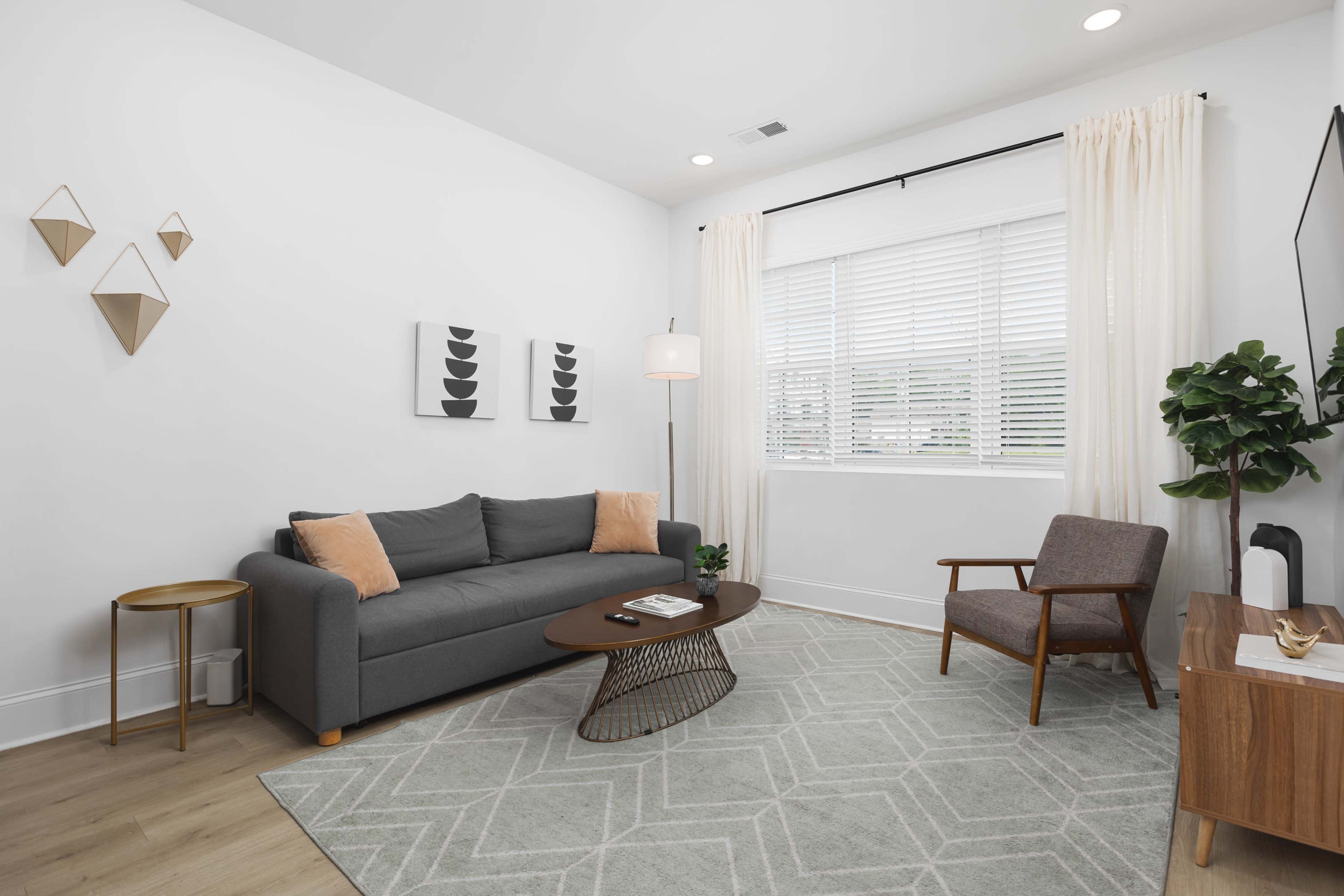 A modern living room features a gray sofa with two cushions, a round coffee table, a chair, and a large window with sheer curtains.