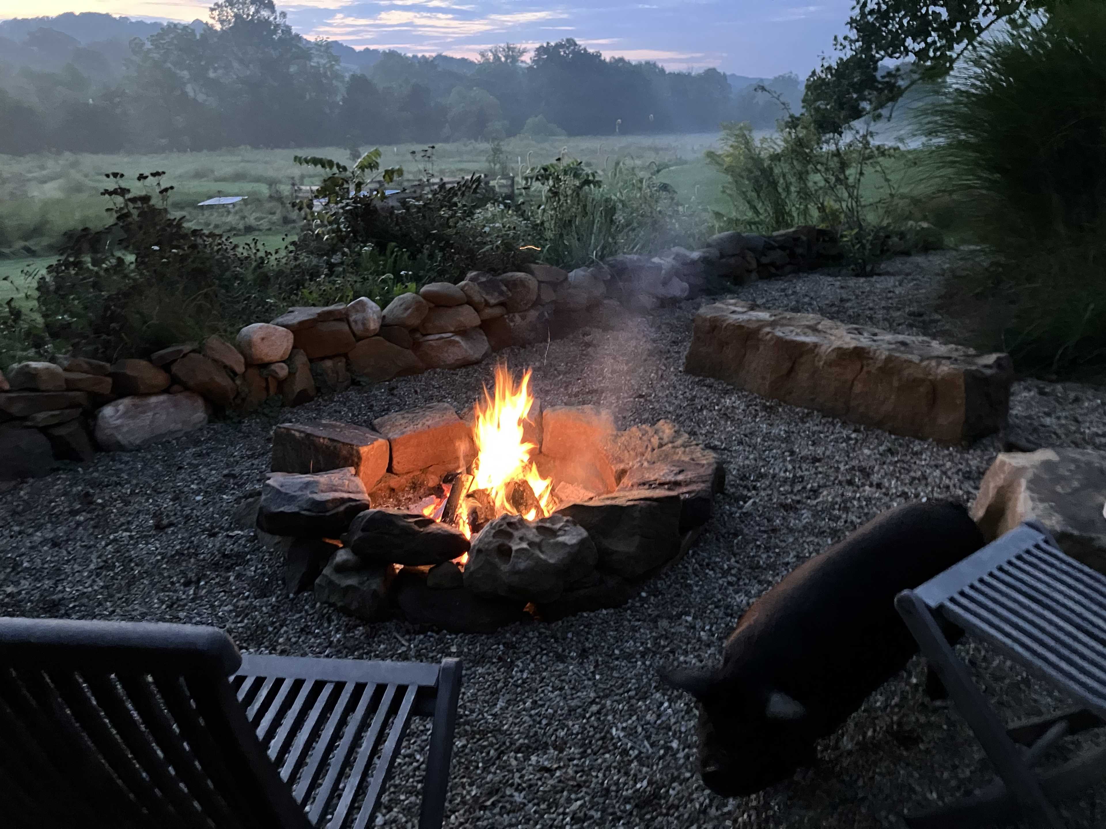 A campfire burns in a stone fire pit surrounded by a gravel area and two chairs, with a pig wandering nearby under a misty sky.