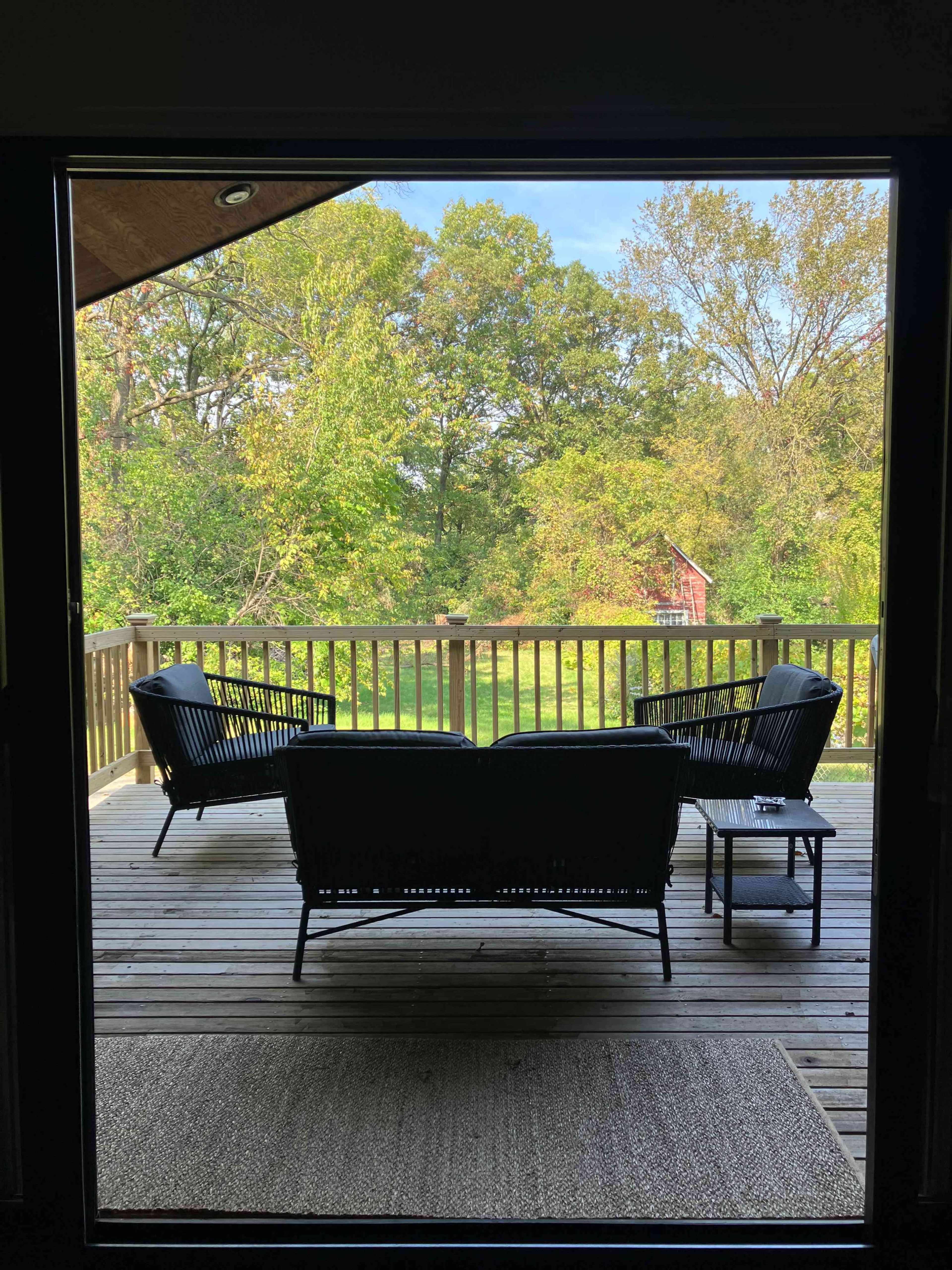 The image shows a covered porch with black wicker furniture overlooking a green landscape filled with trees and a red barn in the distance.