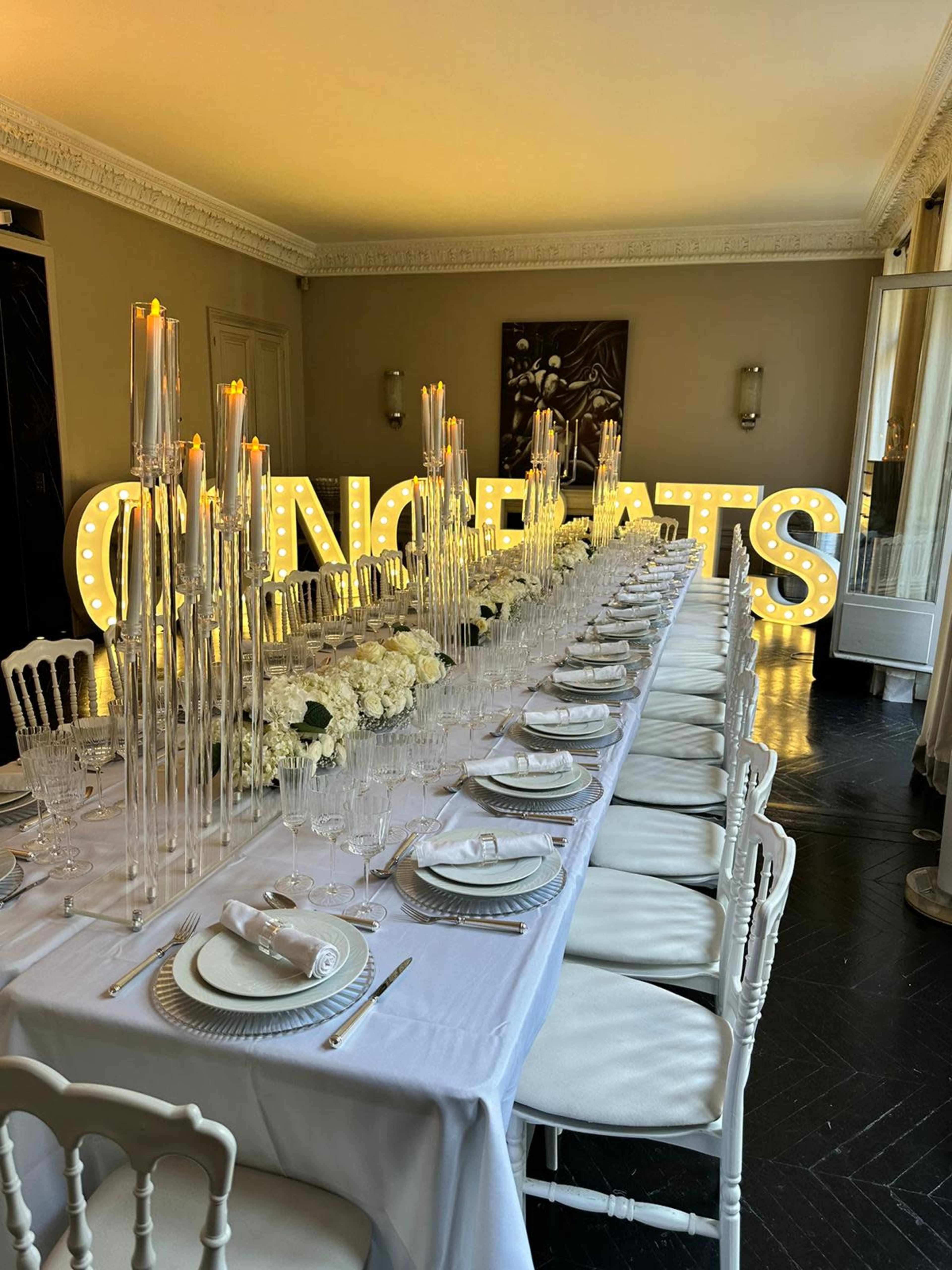 A long, elegantly set dining table is adorned with white flowers and tall candle holders, while a large illuminated "CONGRATS" sign adds a festive touch to the room.