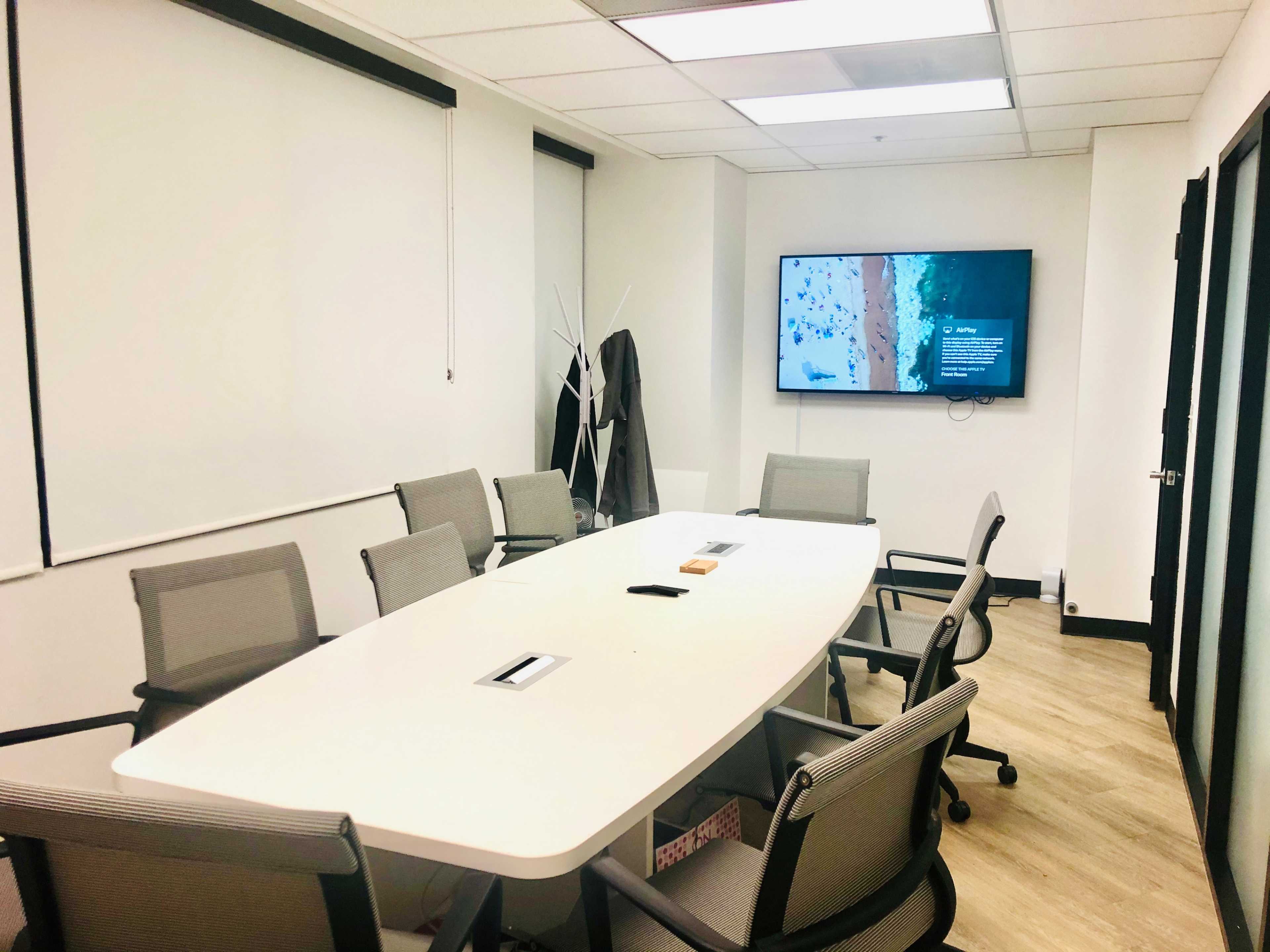 A conference room features a long white table surrounded by several swivel chairs and a wall-mounted television displaying an image.