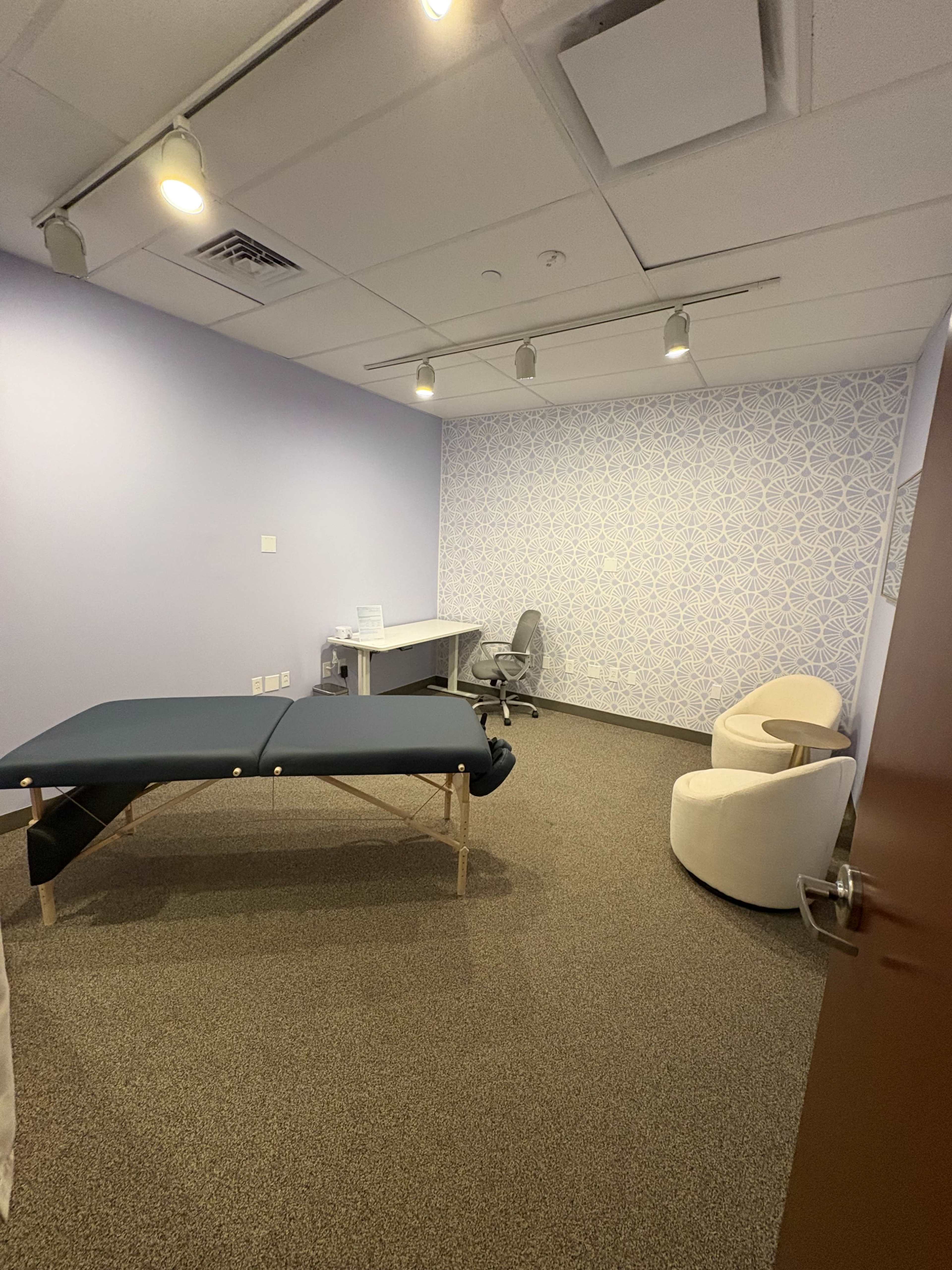 The image shows a clean treatment room featuring a massage table, a desk with a chair, and a circular armchair against a patterned wall.